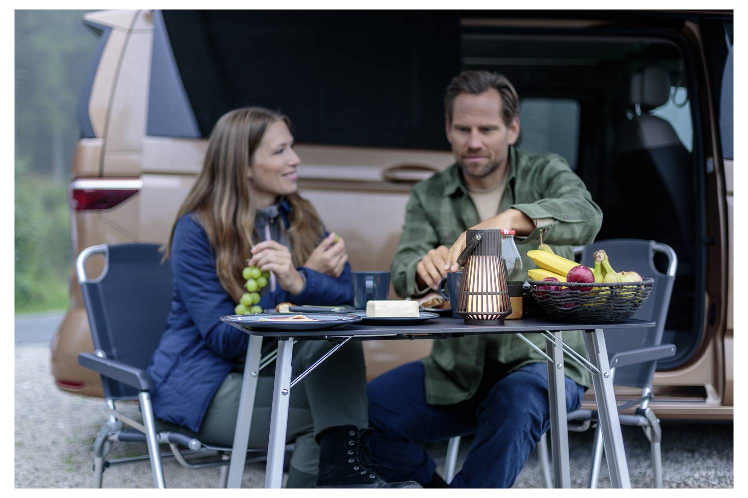 Ein Mann und eine Frau genießen Snacks auf einem Campingplatz und sitzen auf Klappstuhl an einem Lieferwagen. Ein Obstkorb und eine Laterne stehen auf dem Tisch.