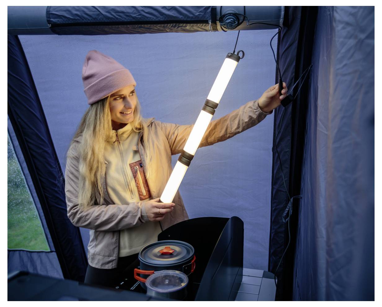 Eine Person in einer Beanie stellt eine helle, vertikale Campinglaterne in einem Zelt auf und beleuchtet die Gegend mit sichtbarer Kochausrüstung in der Nähe.