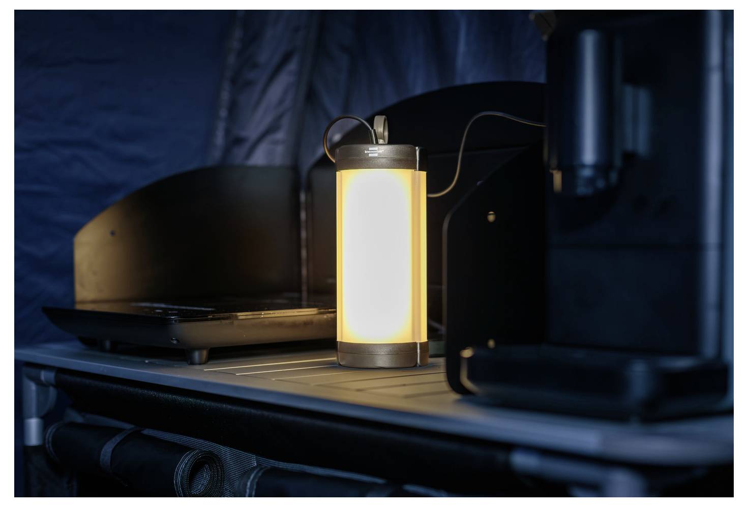 Eine beleuchtete Laterne, die auf einem Tisch in einem Zelt steht, beleuchtet einen Laptop und die umgebende Ausrüstung und deutet auf eine Campingumgebung hin.