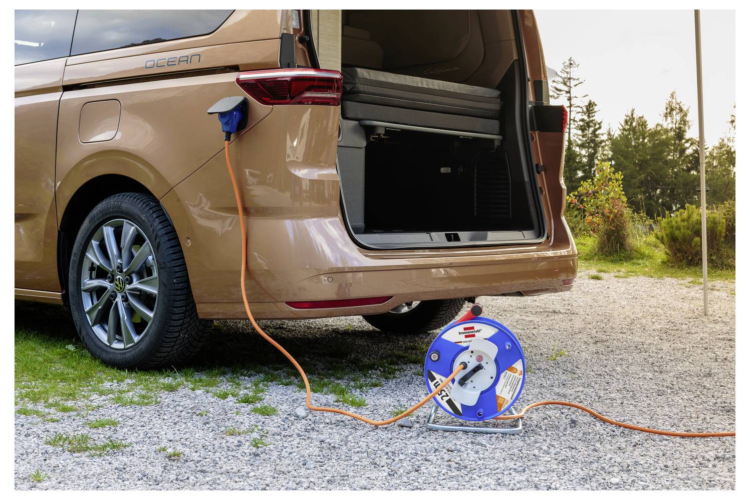 Elektrischer Minivan an ein Ladekabel angeschlossen im Freien, mit einer tragbaren Kabelrolle auf Kies neben der offenen hinteren Heckklappe.