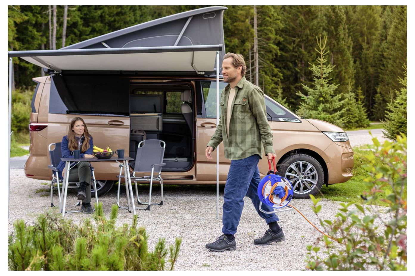 Ein Mann, der ein aufgerolltes orangefarbenes Kabel trägt, geht neben einem beigen Wohnmobil mit Hochdach vorbei, während eine Frau an einem Tisch davor in einem bewaldeten Campingplatz sitzt.
