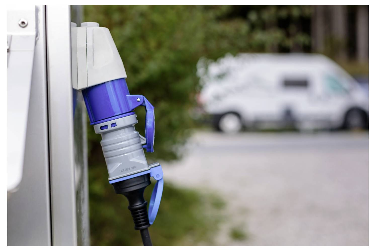 Eine Nahaufnahme eines Ladesteckers für Elektrofahrzeuge, der in eine Steckdose eingesteckt ist, mit einem unscharfen Wohnmobil im Hintergrund auf einer Straße.