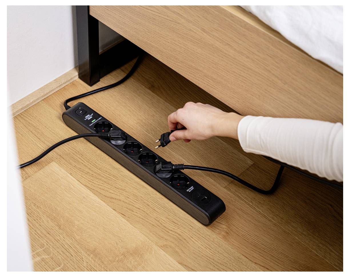 Eine Hand steckt ein Kabel in eine schwarze Steckerleiste auf einem Holzboden neben einem Bett, wobei der Fokus auf Stromnutzung und Konnektivität liegt.