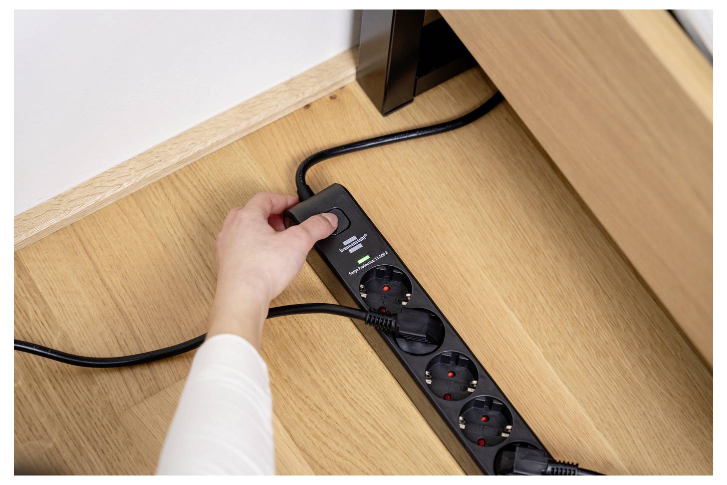 Eine Hand steckt einen Stecker in eine schwarze Steckdosenleiste auf einem Holzboden unter einem Schreibtisch.