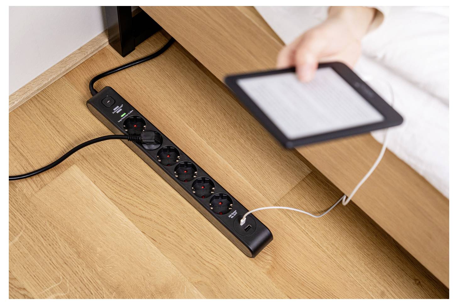 Eine Person lädt einen E-Reader mit einem weißen Kabel auf, das in eine schwarze Steckdosenleiste gesteckt ist, die neben einem Holzschreibtisch angebracht ist.