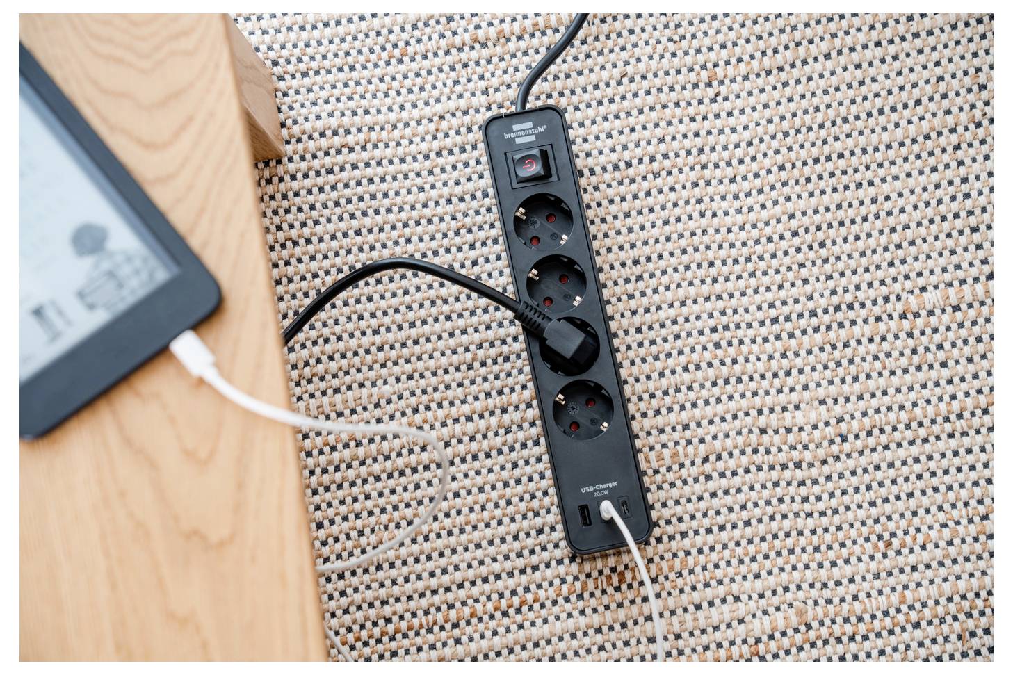 Mehrfachsteckdose auf einem gewebten Teppich neben einem Holztisch, mit mehreren eingesteckten Kabeln, was darauf hinweist, dass elektronische Geräte aufgeladen werden.