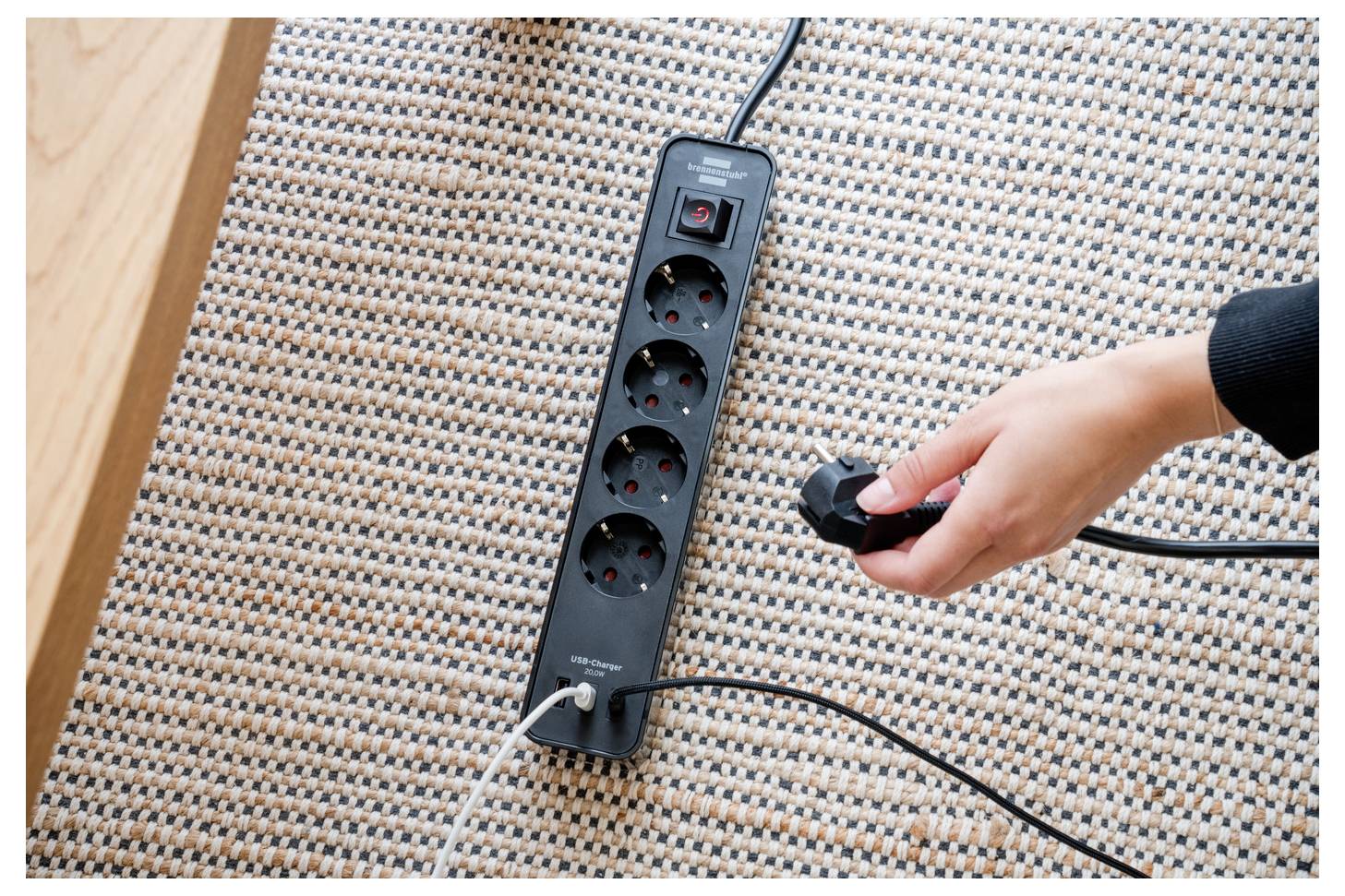 Die Hand einer Person steckt ein Kabel in eine schwarze Steckdosenleiste mit vier Anschlüssen, die auf einem strukturierten Teppich platziert ist.