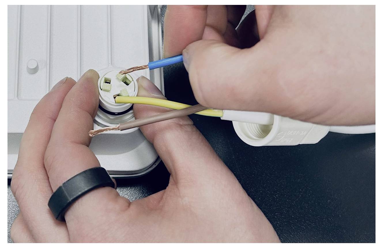 Hände verdrahten einen dreipoligen Stecker und verbinden blaue, braune und gelb-grüne Drähte mit einer Steckdosenklemme.