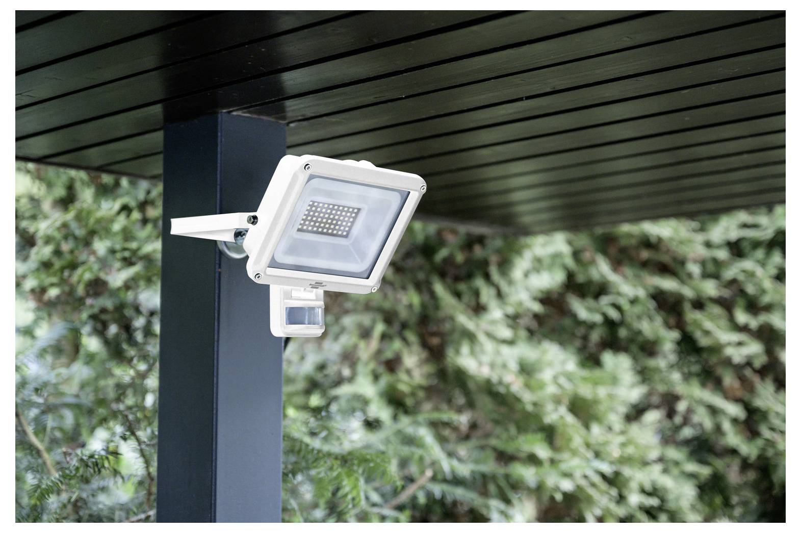 Außenstrahler mit Bewegungsmelder an einer Terrassendachstütze montiert, umgeben von grünem Laub.