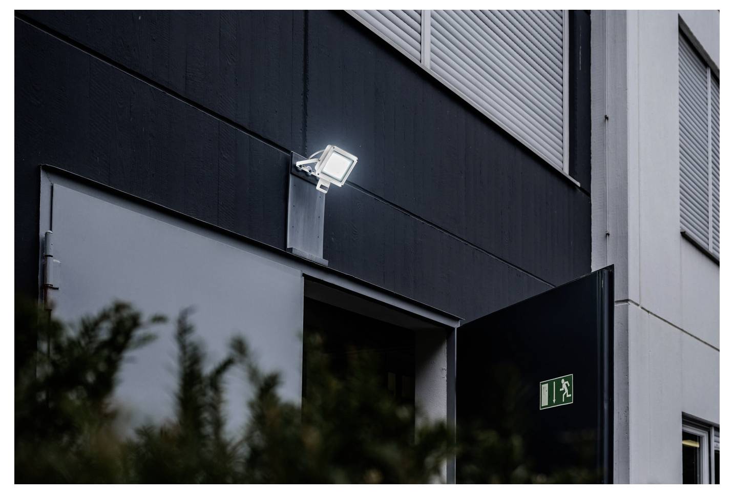 Eine offene Tür mit einem Notausgangsschild darüber, beleuchtet von einem externen Flutlicht an einer dunklen Gebäudefassade. Büsche im Vordergrund.