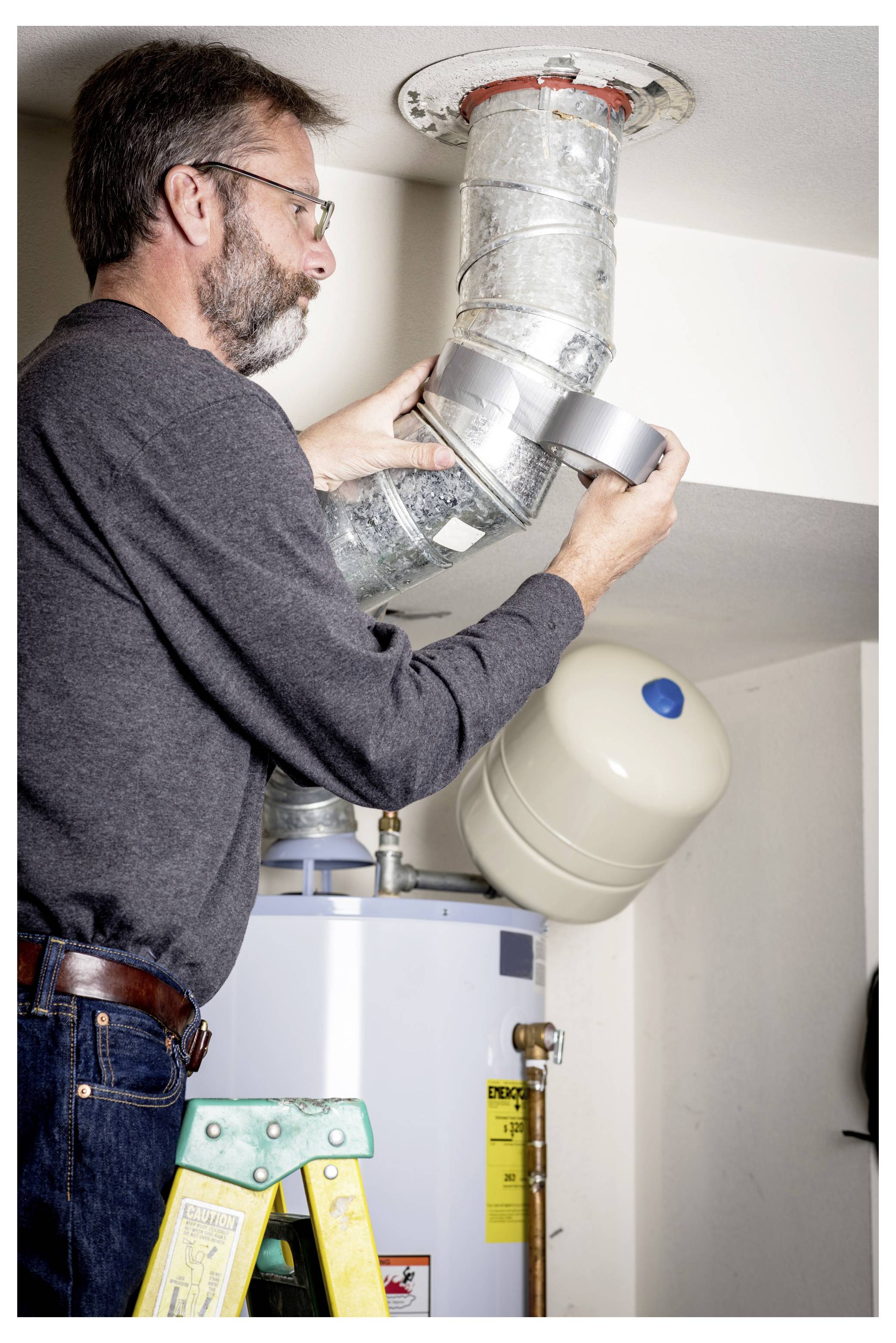 Ein Mann installiert Rohrleitungen an einem Warmwasserspeicher in einem Heizungsraum, wobei er eine Stehleiter verwendet.