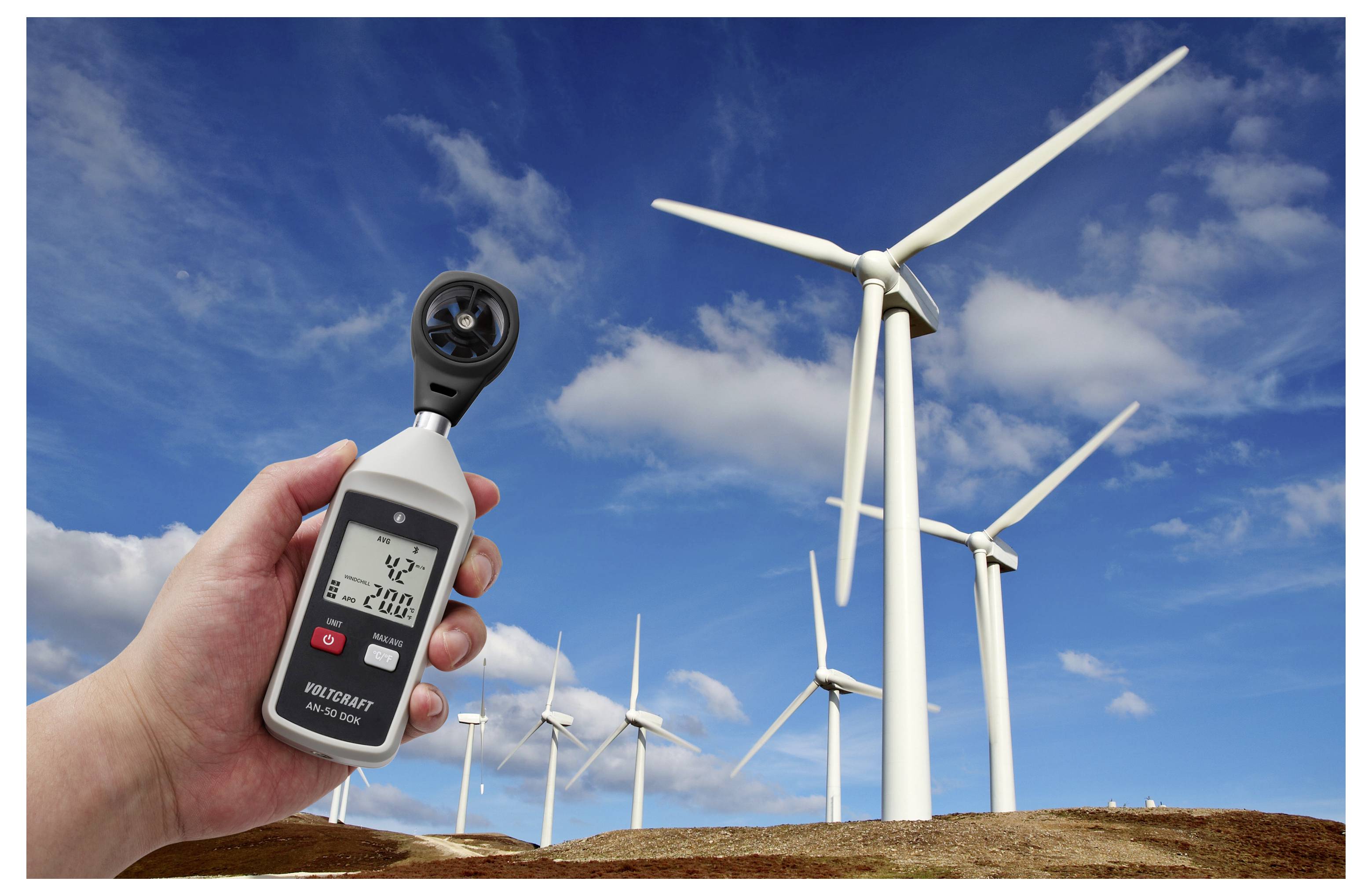 Hand hält ein digitales Anemometer, das die Windgeschwindigkeit vor Windkraftanlagen auf einem Windpark unter blauem Himmel misst.