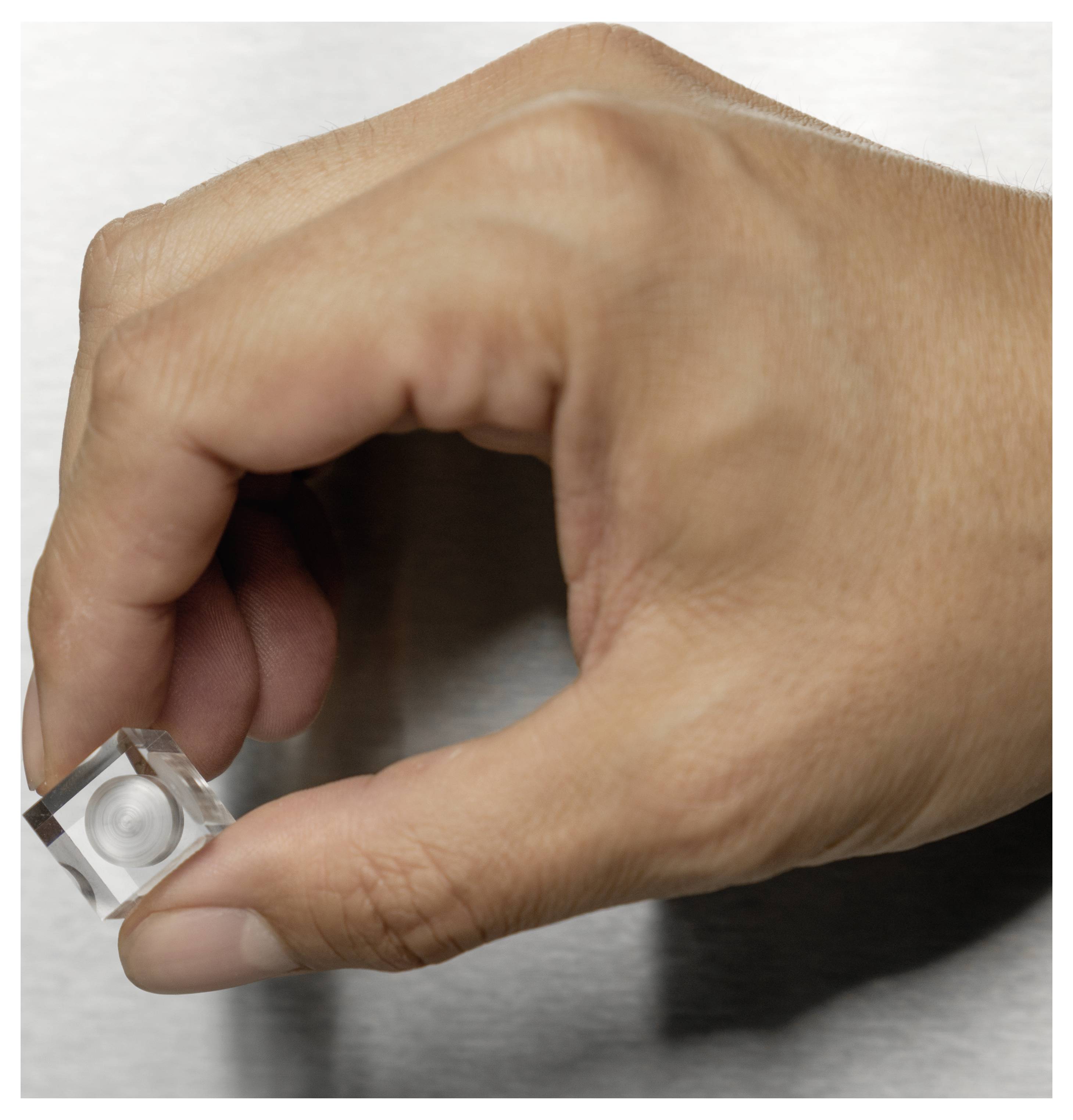 Eine Hand, die einen kleinen, transparenten Würfel mit einer kreisförmigen Vertiefung hält, vor einem einfachen Hintergrund positioniert.