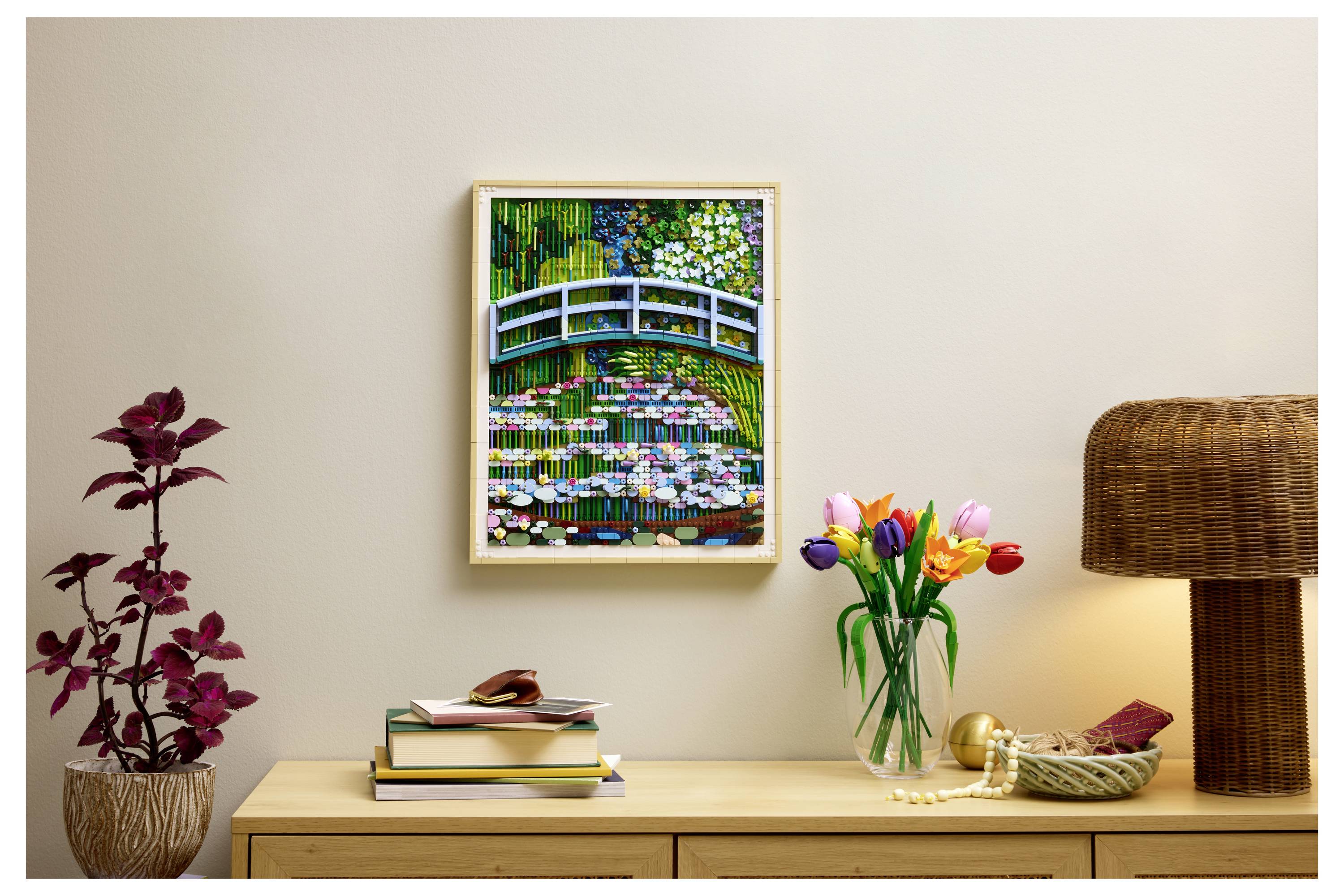 Ein gerahmtes Gemälde einer Brücke über Seerosen hängt an einer Wand über einer Holzkommode mit Büchern, einer Vase mit Tulpen und einer geflochtenen Lampe.