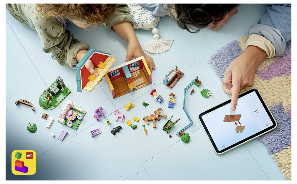 Zwei Personen bauen ein LEGO-Haus und spielen mit verstreuten Teilen, während sie auf ein Tablet zeigen, das ein Spielzeugboot auf dem Bildschirm anzeigt.