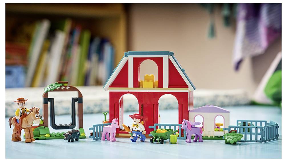 Spielszene mit Spielzeugbauernhof mit einer roten Scheune, rosa Ponys, einer Cowboy-Figur auf einem Pferd und kleinen Zäunen sowie Zubehör auf einer Tischplatte.