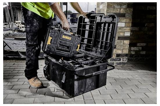 Eine Person in Arbeitsschuhen und einer Sicherheitsweste stellt eine Werkzeugspeicherbox in eine größere Werkzeugkiste auf einer Baustelle mit sichtbarem Mauerwerk.