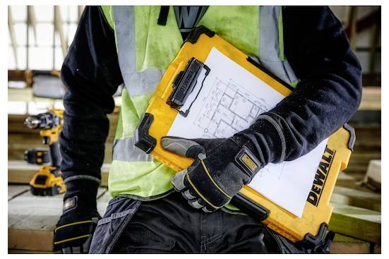 „Person in Arbeitskleidung hält ein gelbes DeWalt-Klemmbrett mit Bauplänen und steht auf einer Baustelle mit Werkzeugen im Hintergrund.