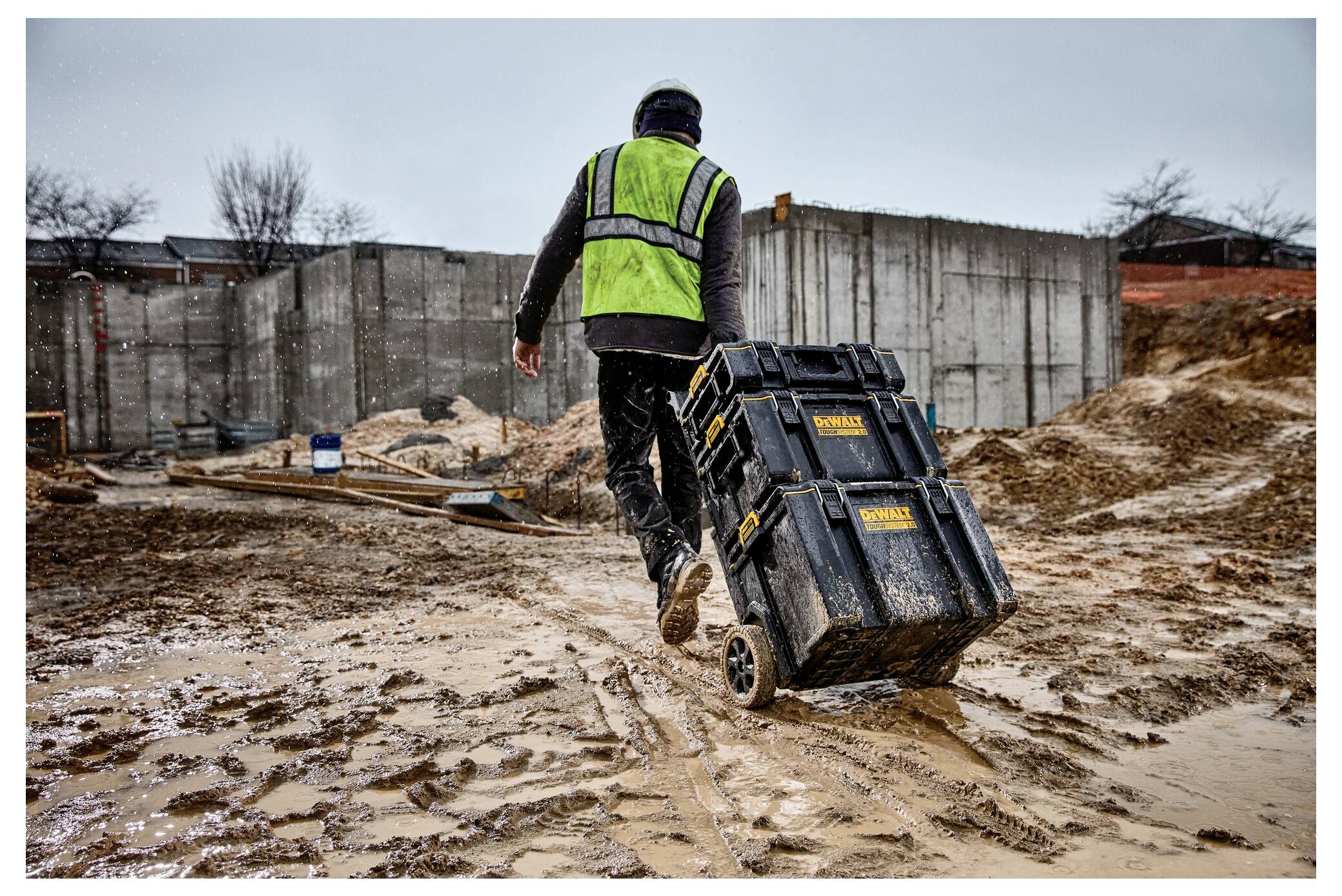 Eine Person in einer grünen Weste und einem Helm zieht einen Werkzeugwagen über eine schlammige Baustelle mit Betonwänden und verstreuten Materialien.