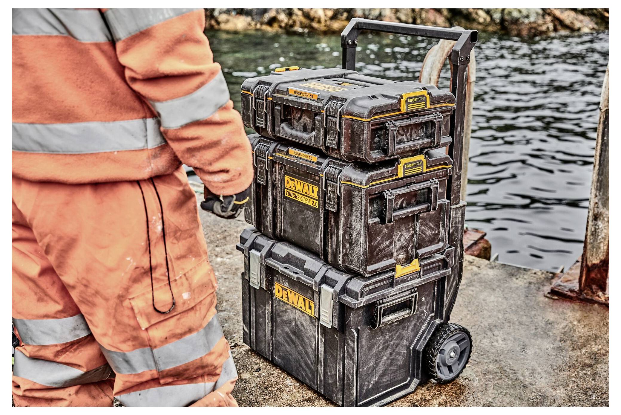 Ein Arbeiter in orangener Kleidung steht neben einem Stapel von DeWalt-Werkzeugkästen am Wasser auf einer Betonoberfläche. Die Einrichtung ist für industrielle Verwendung vorgesehen.