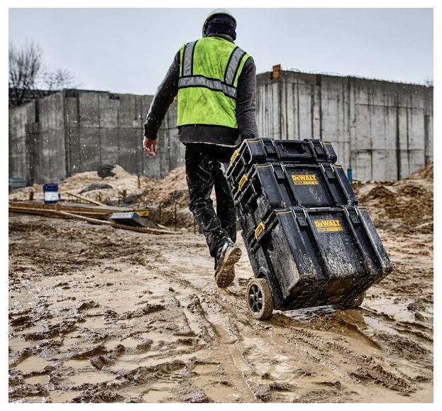 Ein Bauarbeiter in einer grünen Sicherheitsweste zieht eine Werkzeugkiste mit Rädern über eine schlammige Baustelle, mit Betonmauern im Hintergrund.