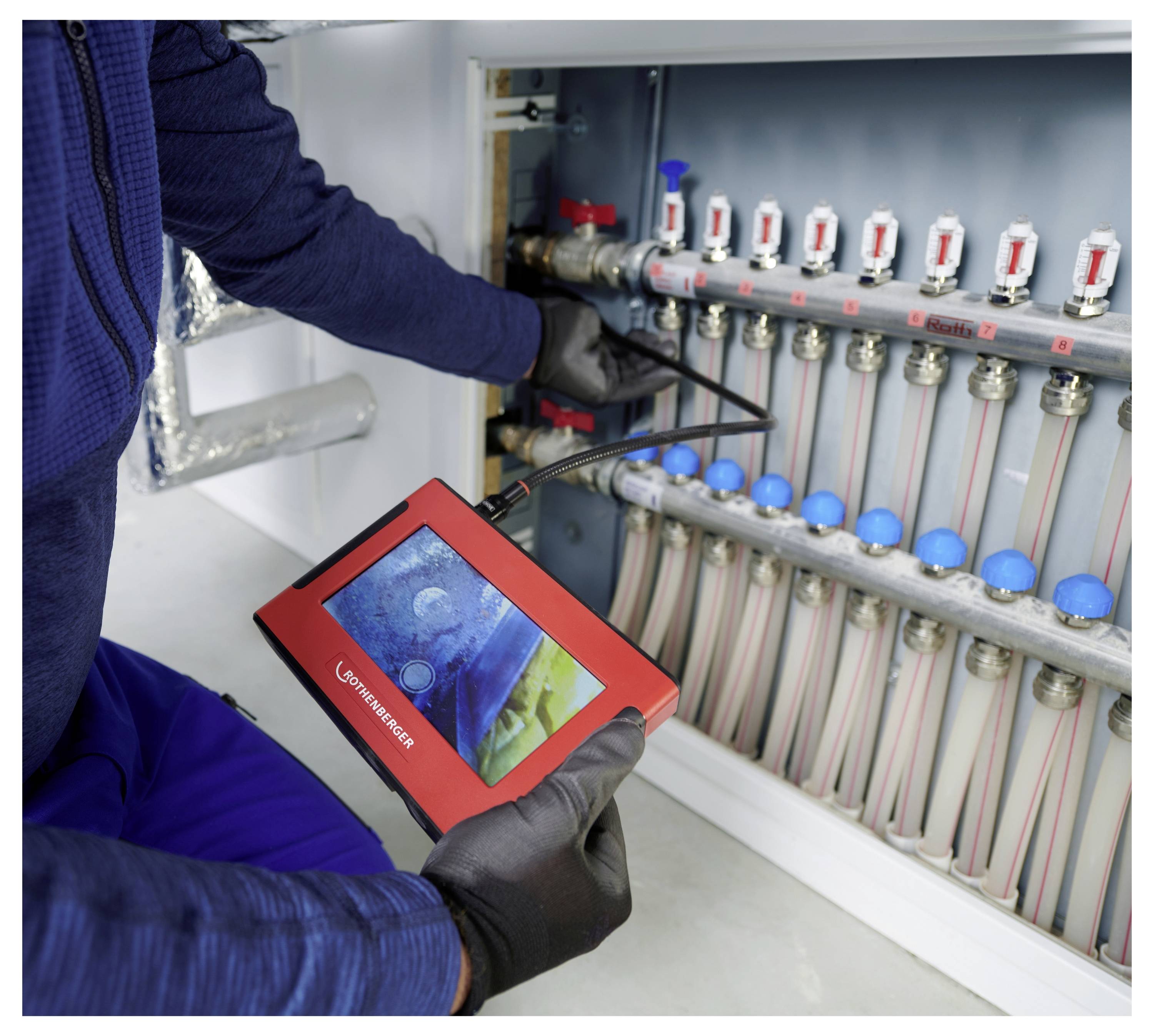 Eine Person in blauem Arbeitsoverall nutzt ein Diagnose-Werkzeug, um eine Reihe von Rohren und Ventilen zu überprüfen, was auf Wartungsarbeiten an einem Heizsystem hindeutet.