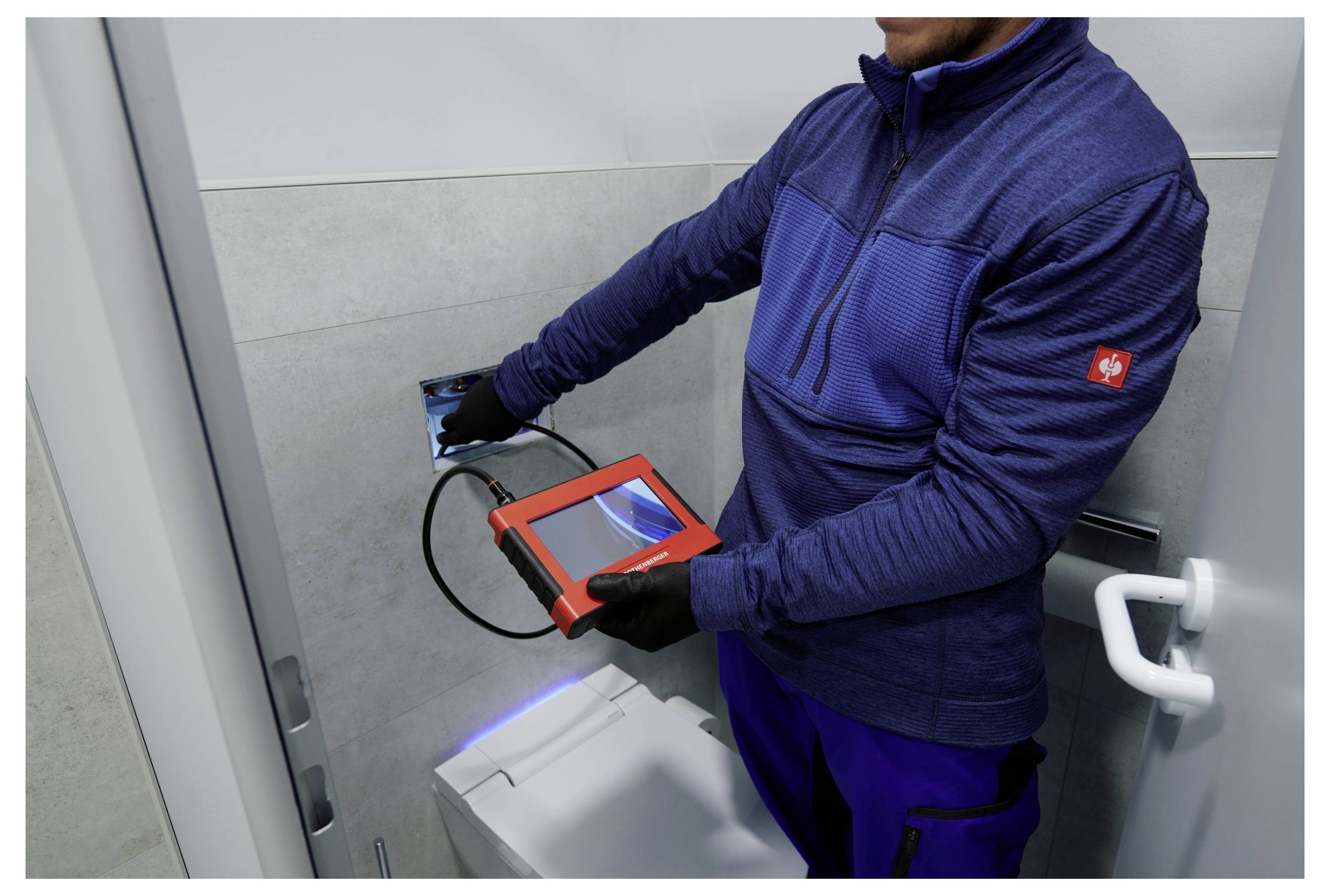 Eine Person in einer blauen Jacke benutzt ein rotes Inspektionsgerät mit einer Kamera, um einen Wandbereich im Badezimmer zu überprüfen.