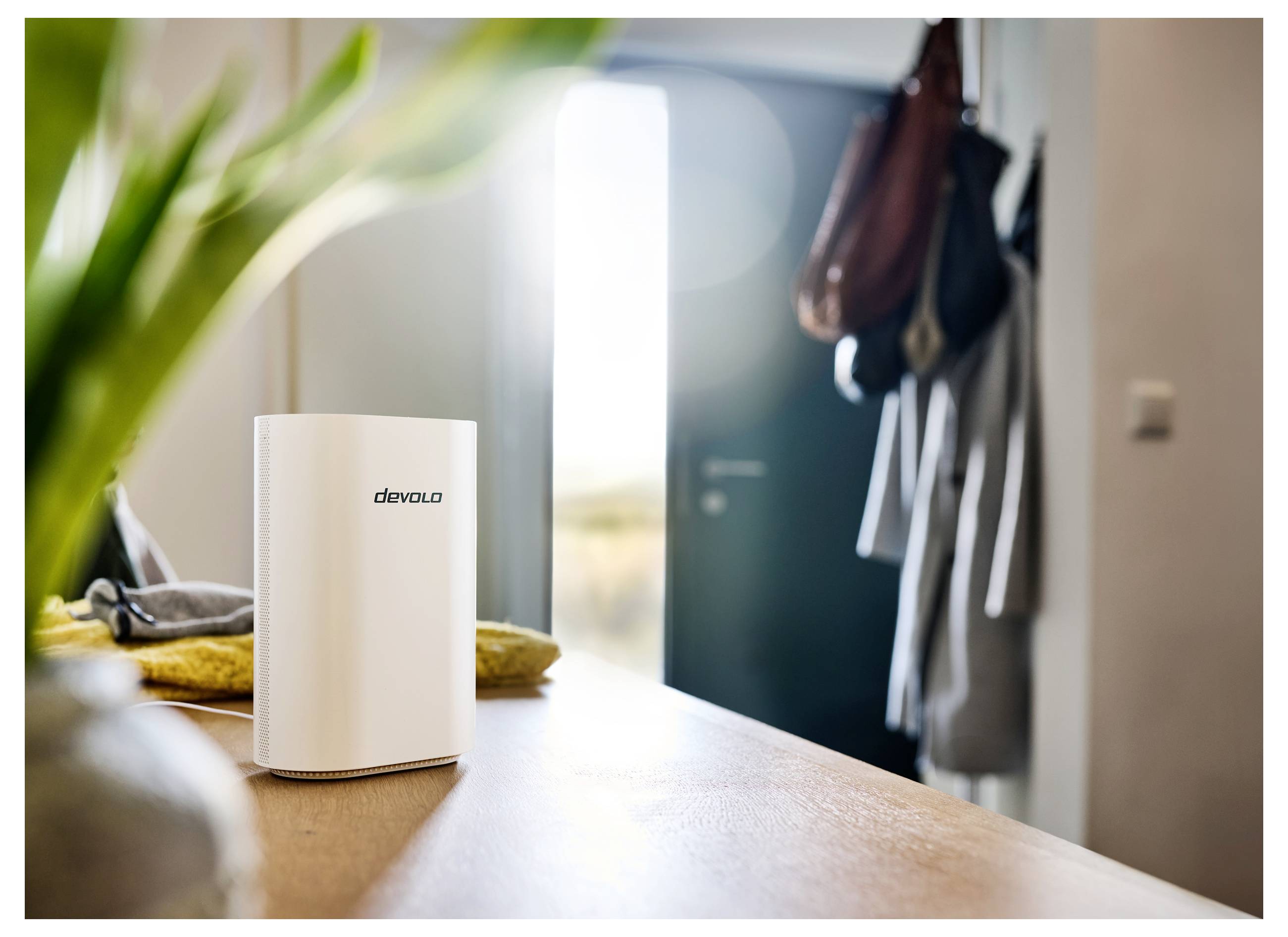 Ein weißer Devolo-Router steht auf einem Tisch in einem gut beleuchteten Raum, mit Blick auf eine teilweise offene Tür und hängende Mäntel im Hintergrund.