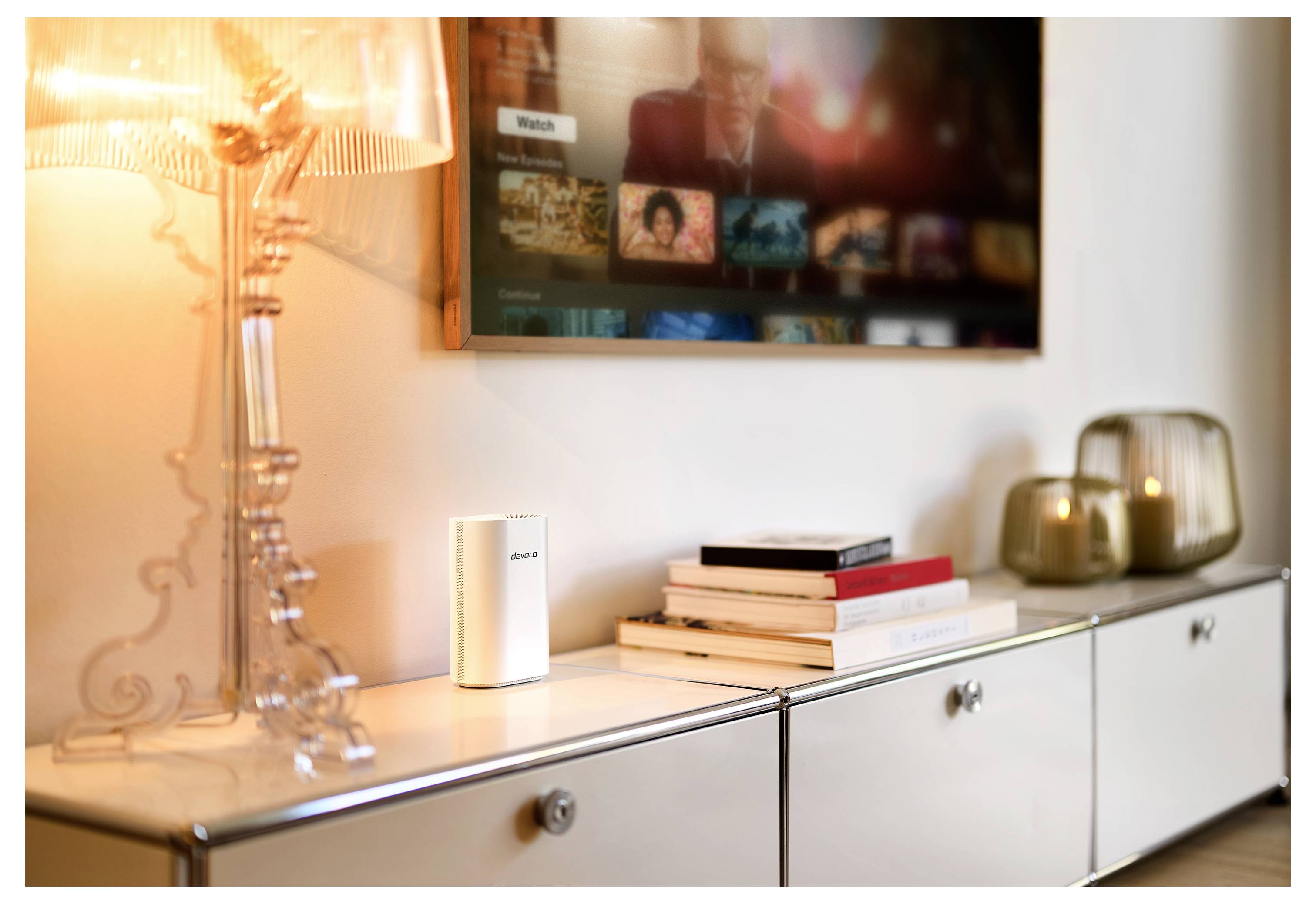 Ein minimalistisches Wohnzimmer mit einer modernen Lampe, einem Bücherstapel und brennenden Kerzen auf einem weißen Schrank; Fernsehbildschirm im Hintergrund.