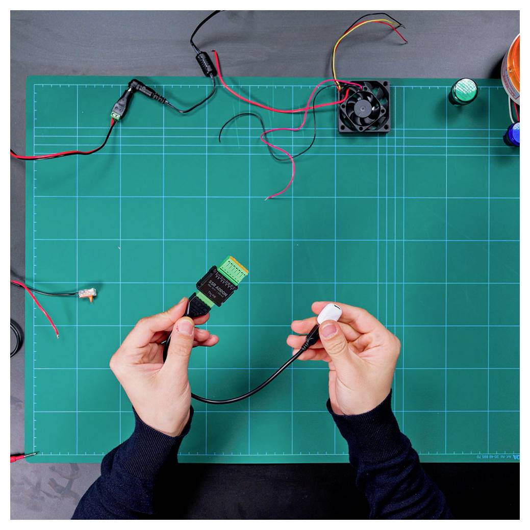 Hände halten elektronische Stecker über eine grüne Arbeitsmatte mit Drähten, einem Lüfter und kleinen Komponenten, was auf Elektronik-Montage hindeutet.