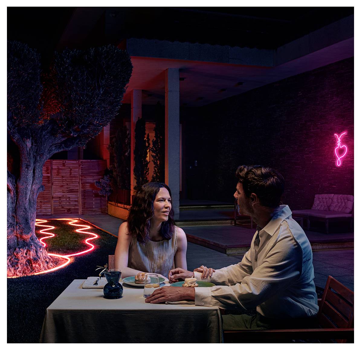 Ein Paar sitzt an einem Tisch im Freien eines Restaurants unter abendlichen Lichtern und unterhält sich. Ein Neonlicht schmückt den Hintergrund.
