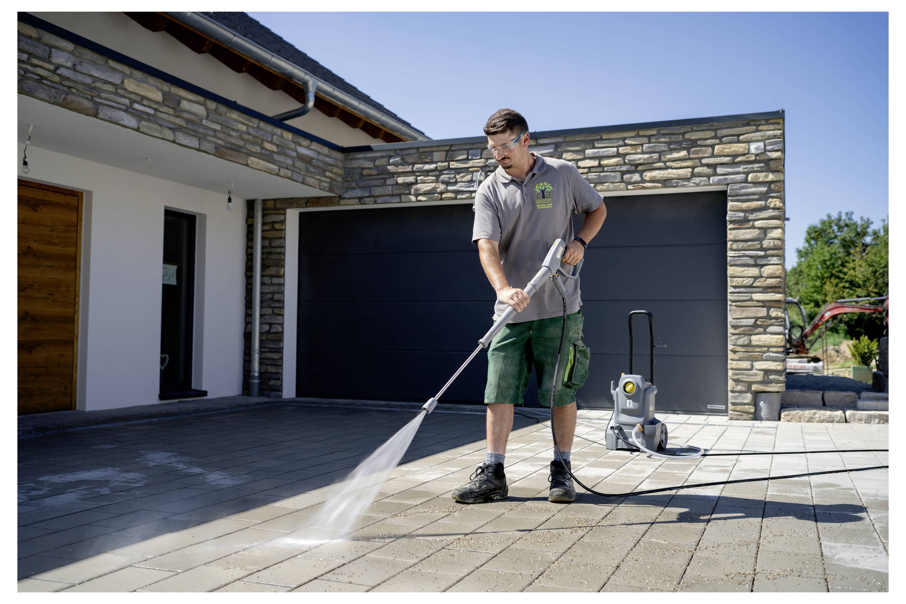 Eine Person benutzt einen Hochdruckreiniger, um eine gepflasterte Auffahrt vor einer Garage zu reinigen, trägt zwanglose Arbeitskleidung und konzentriert sich auf die Aufgabe.