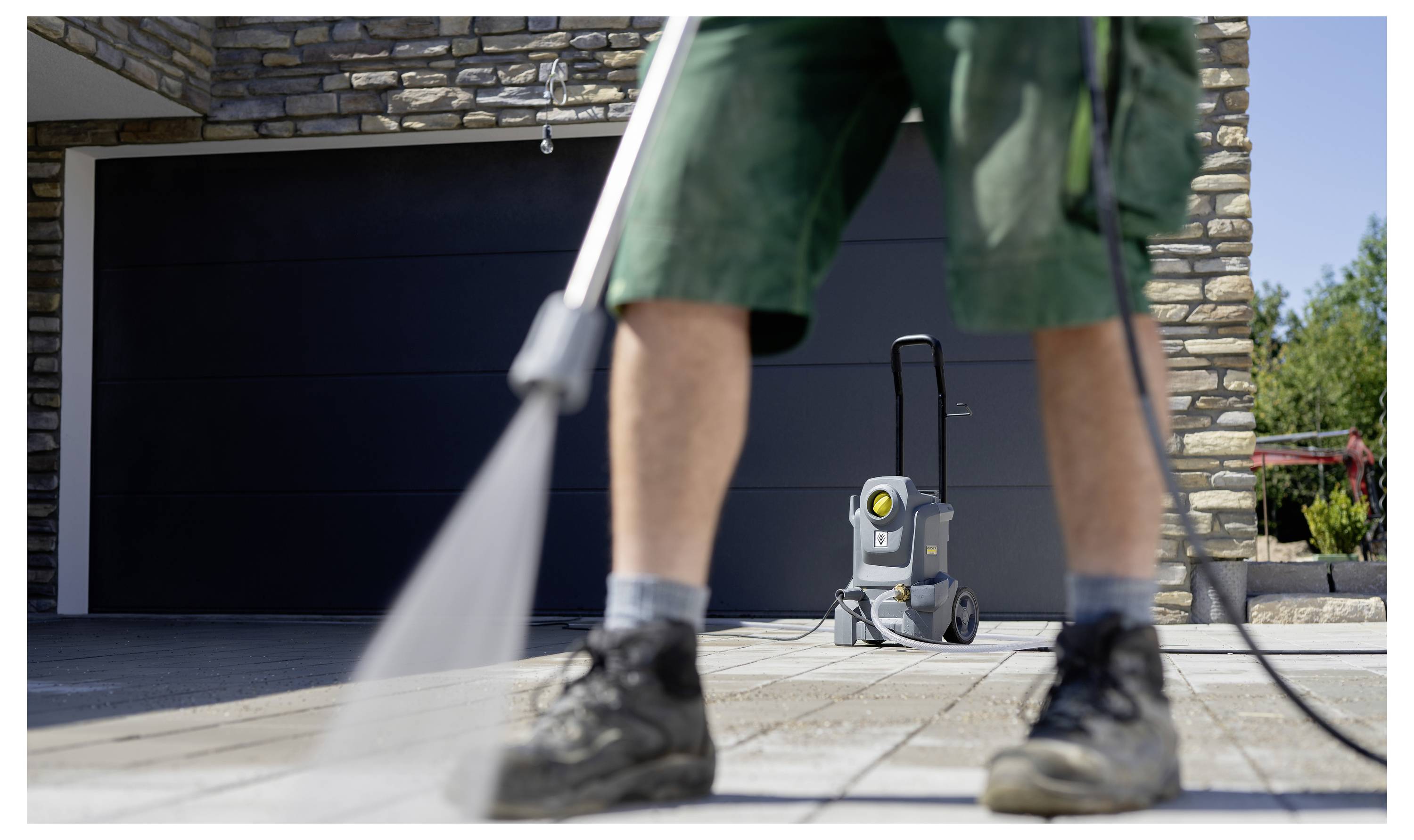 Eine Person in Shorts benutzt einen Hochdruckreiniger, um eine Steineinfahrt vor einem Backsteinhaus mit sichtbarer Garagentür zu reinigen.
