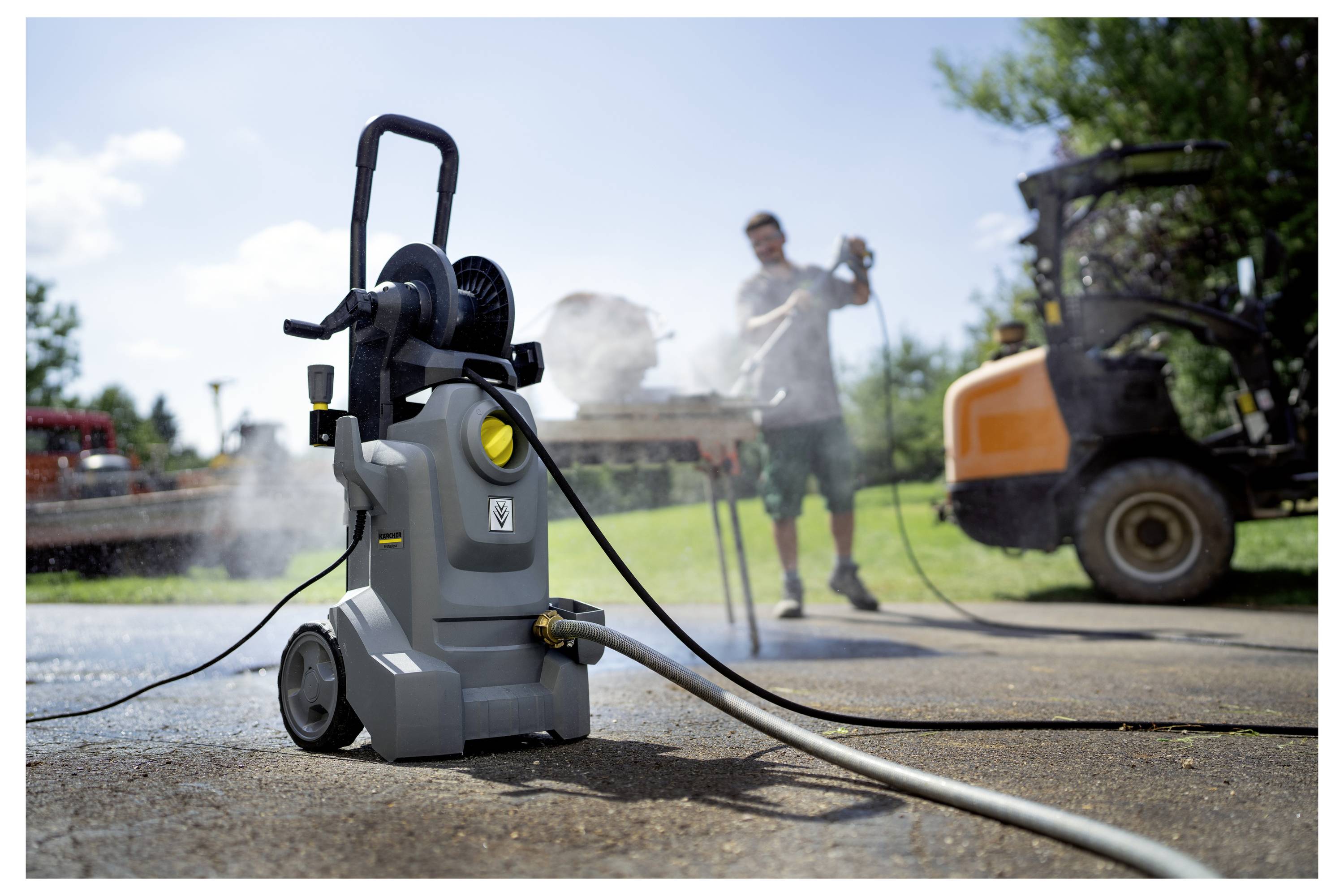 Eine Person benutzt einen Hochdruckreiniger auf einer Einfahrt, mit einem Schlauch, der daran befestigt ist. Ein Fahrzeug und Bäume sind im Hintergrund sichtbar.