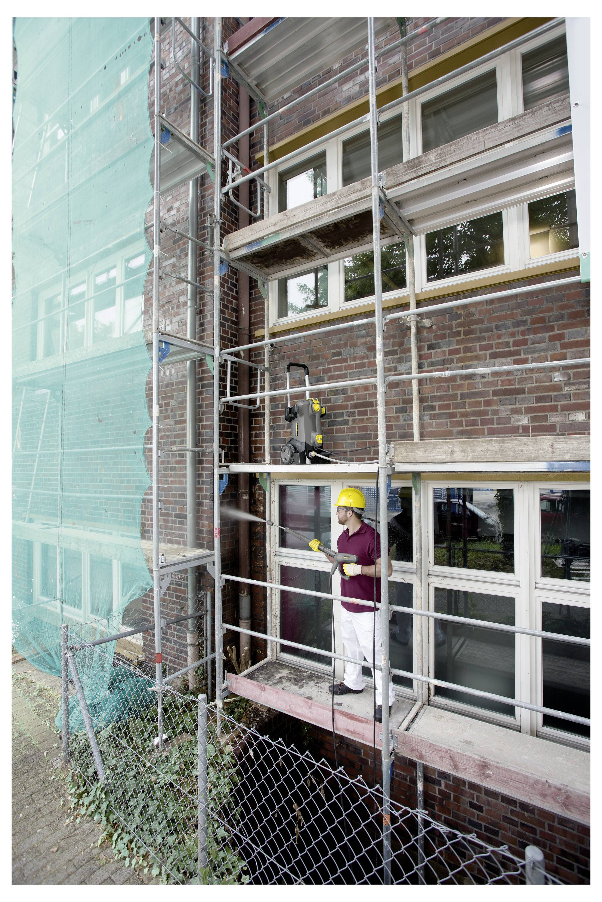 Ein Bauarbeiter in einem gelben Schutzhelm steht auf Gerüsten außerhalb eines Backsteingebäudes, trägt Schutzausrüstung, mit sichtbaren Werkzeugen.