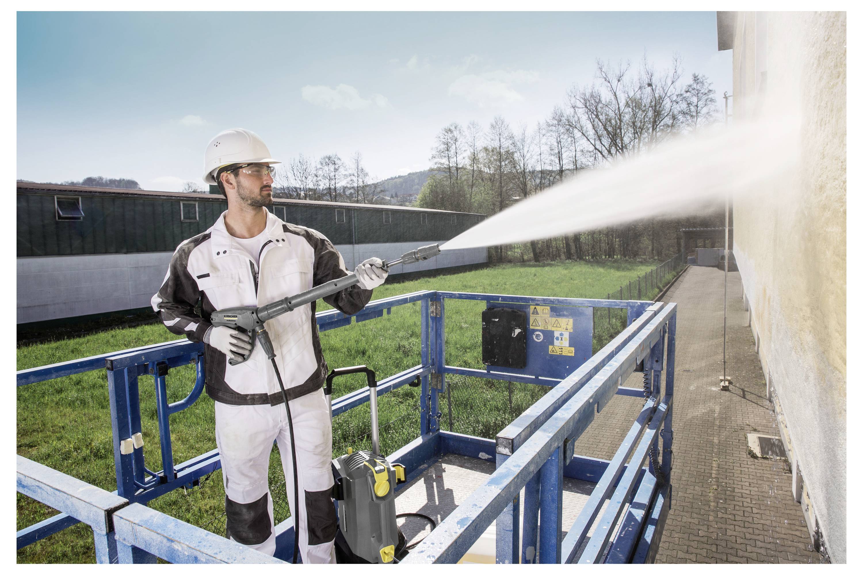Ein Arbeiter in Schutzausrüstung reinigt mit einem Hochdruckreiniger die Außenfassade eines Gebäudes und steht dabei auf einer blauen Hubarbeitsbühne im Freien.
