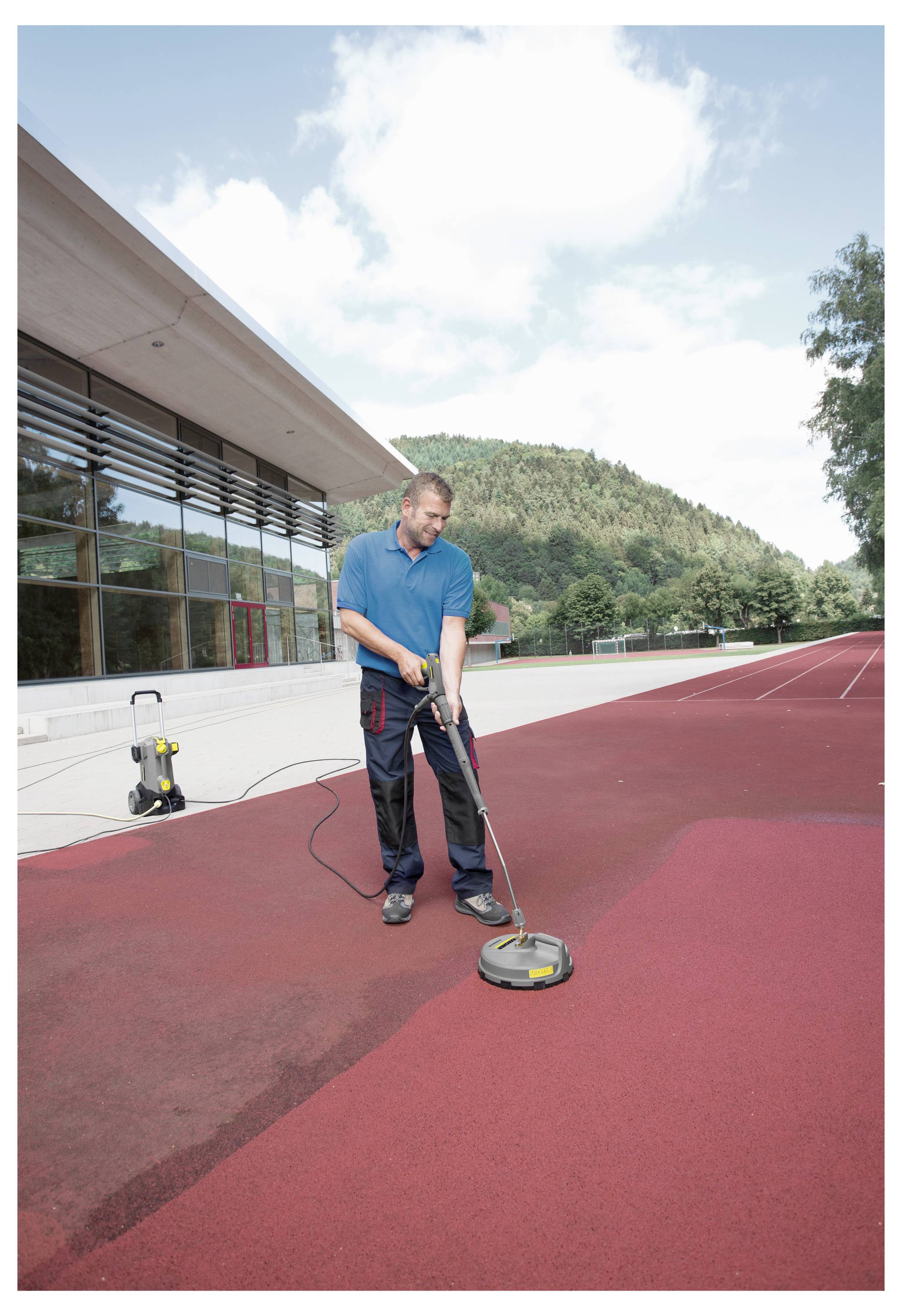 Eine Person, die eine rote Außensportanlage mit einem Hochdruckreiniger reinigt, neben einem modernen Gebäude und Bäumen im Hintergrund.