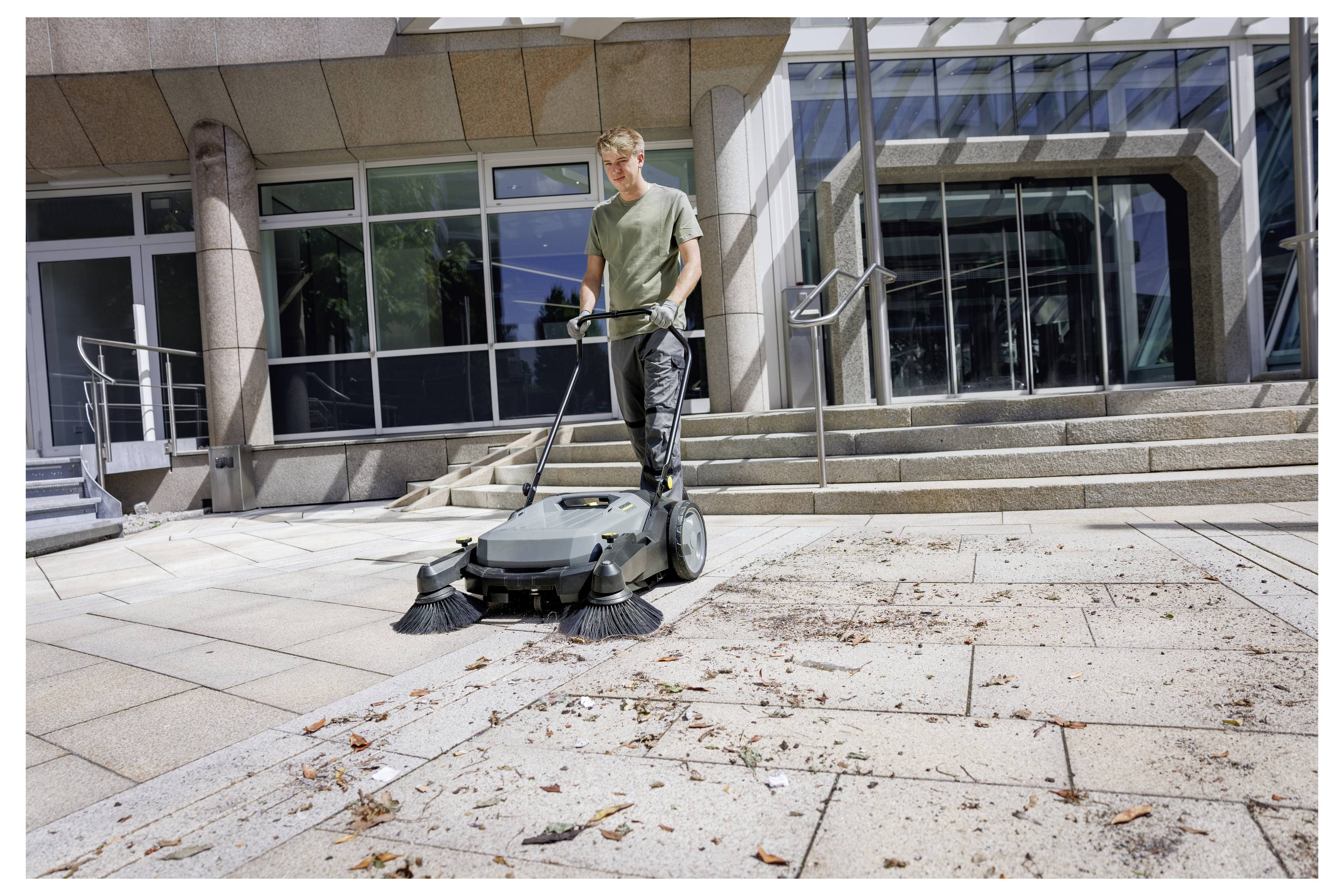 Eine Person nutzt einen Outdoor-Besen, um Schutt auf einem großen, gefliesten Platz vor einem modernen Gebäude mit Glas- und Metallarchitektur zu reinigen.