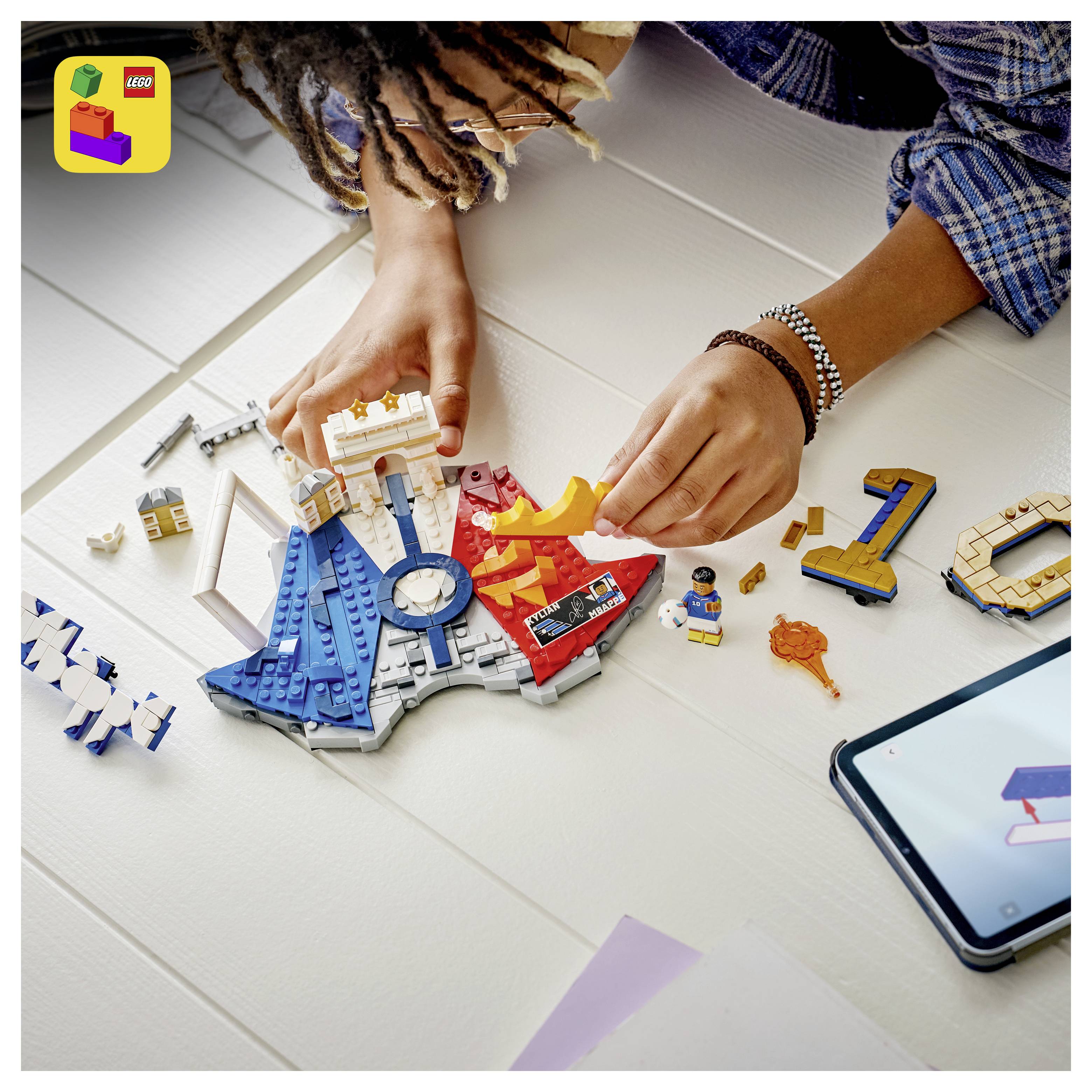 Person, die ein buntes LEGO-ähnliches Modell zusammenbaut, mit einem Smartphone in der Nähe, mit lose verstreuten Teilen und einer Minifigur auf einem Tisch.