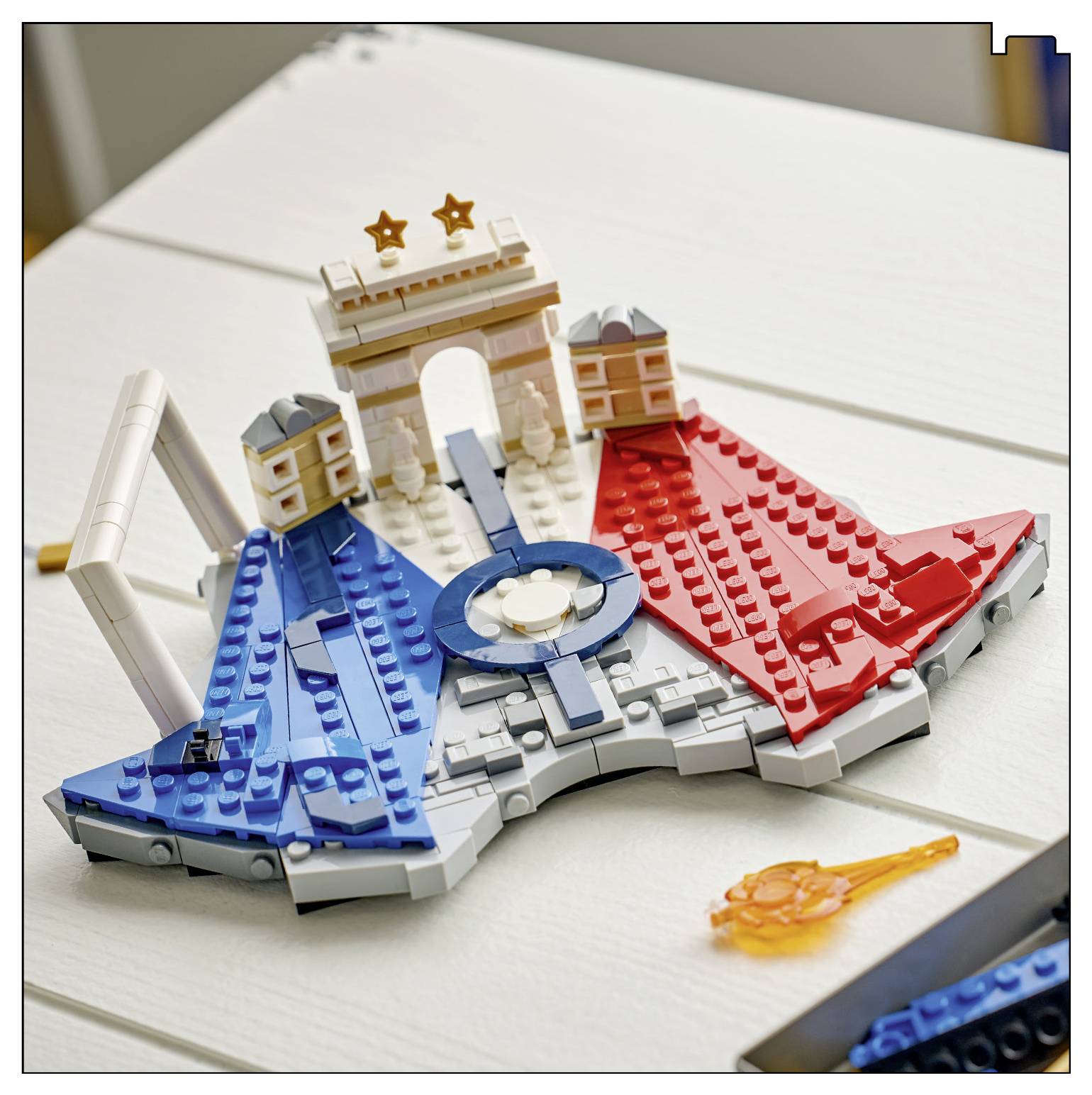 LEGO-artiges Modell eines Stadions oder einer Arena mit blauen und roten Abschnitten, einem zentralen kreisförmigen Element und einem Bogen mit Sternen auf einem weißen Tisch.