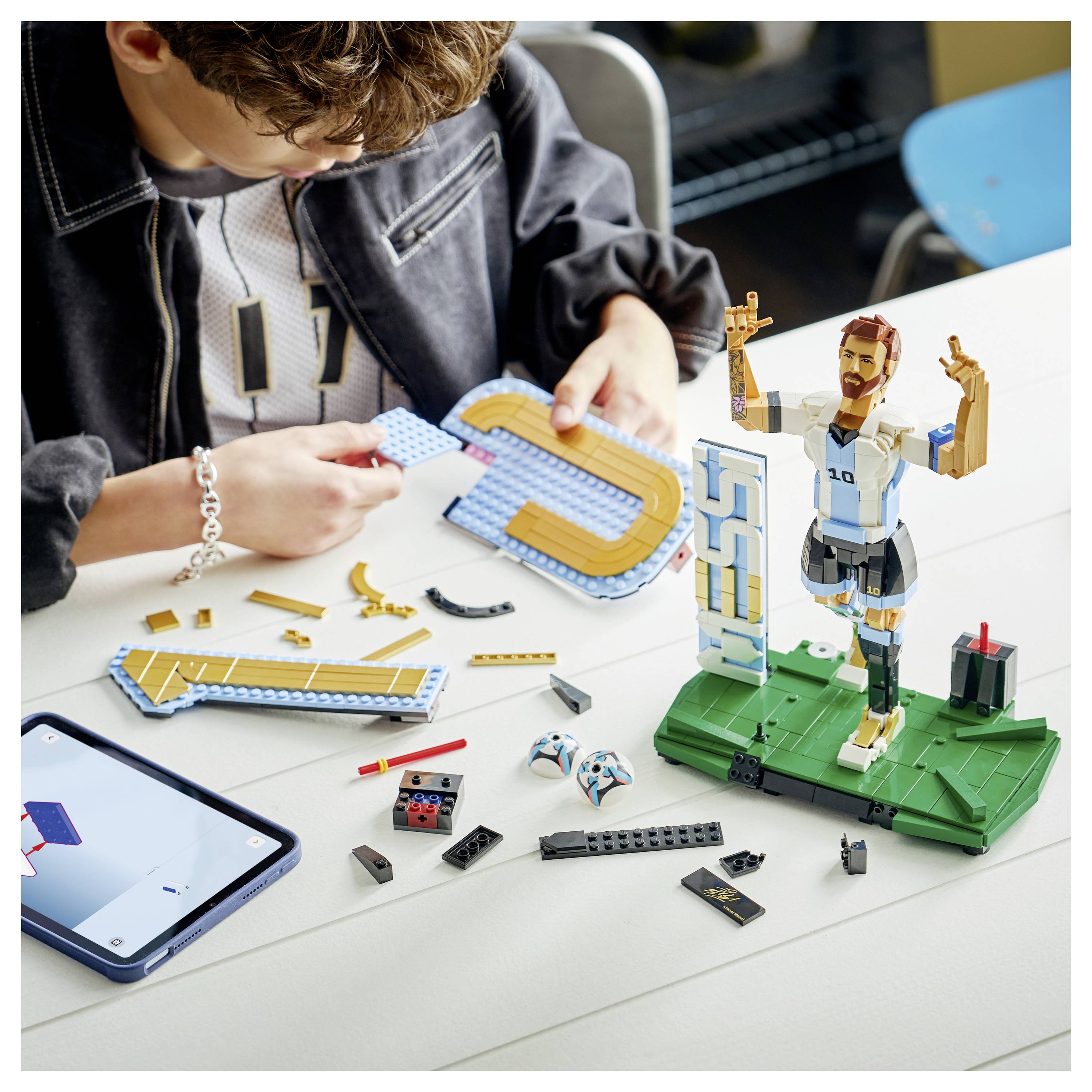 Ein Kind baut eine LEGO-Basketballszene zusammen, mit einer Spielerfigur und einer Korbanzeige auf einem Tisch neben verstreuten Teilen und einem Telefon.