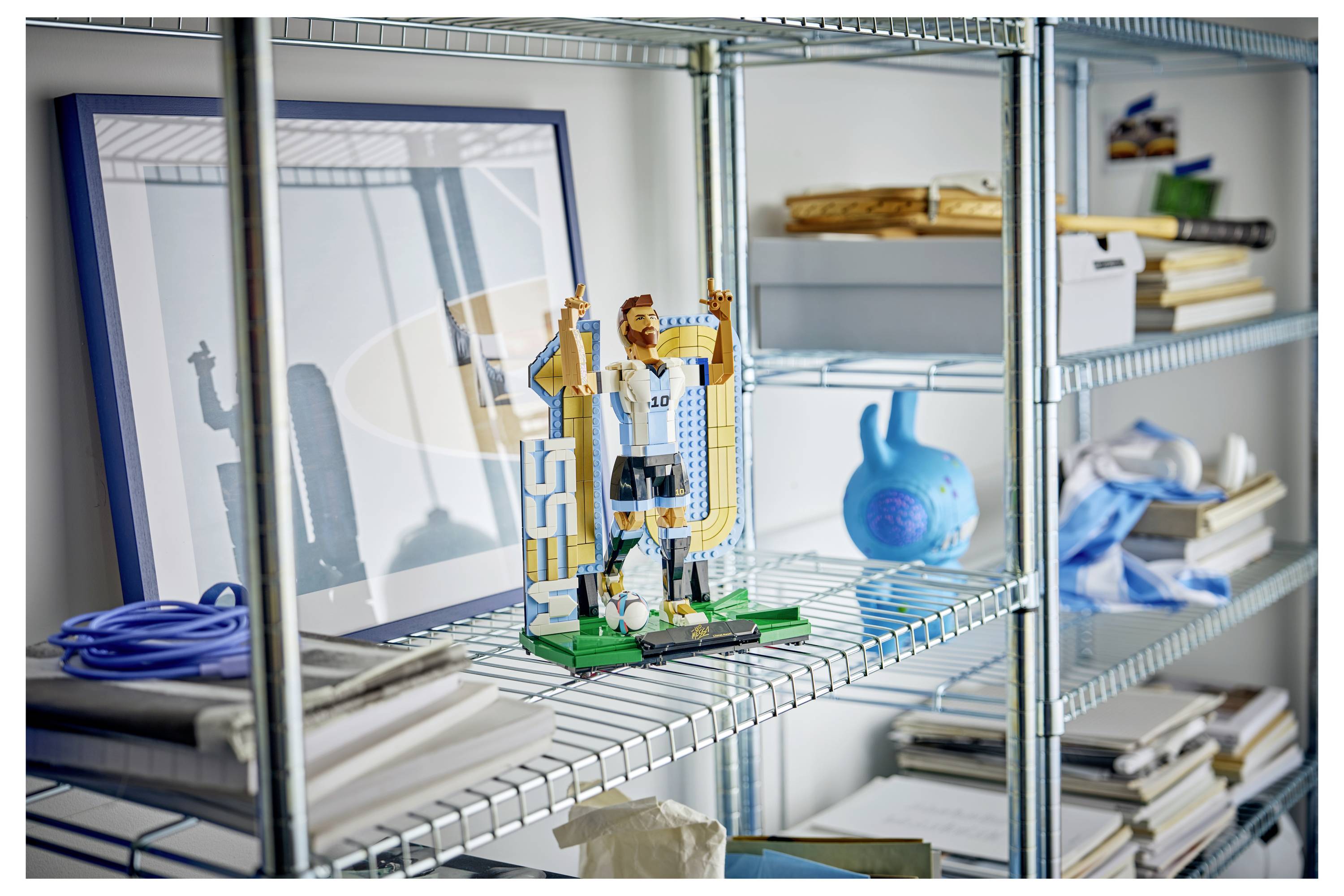 Eine kleine LEGO-ähnliche Figur, die zwei goldene Pokale hält, sitzt auf einem Drahtregal in einem Lagerraum, in der Nähe einer blauen Vase, Bücher und gerahmte Kunstwerke.