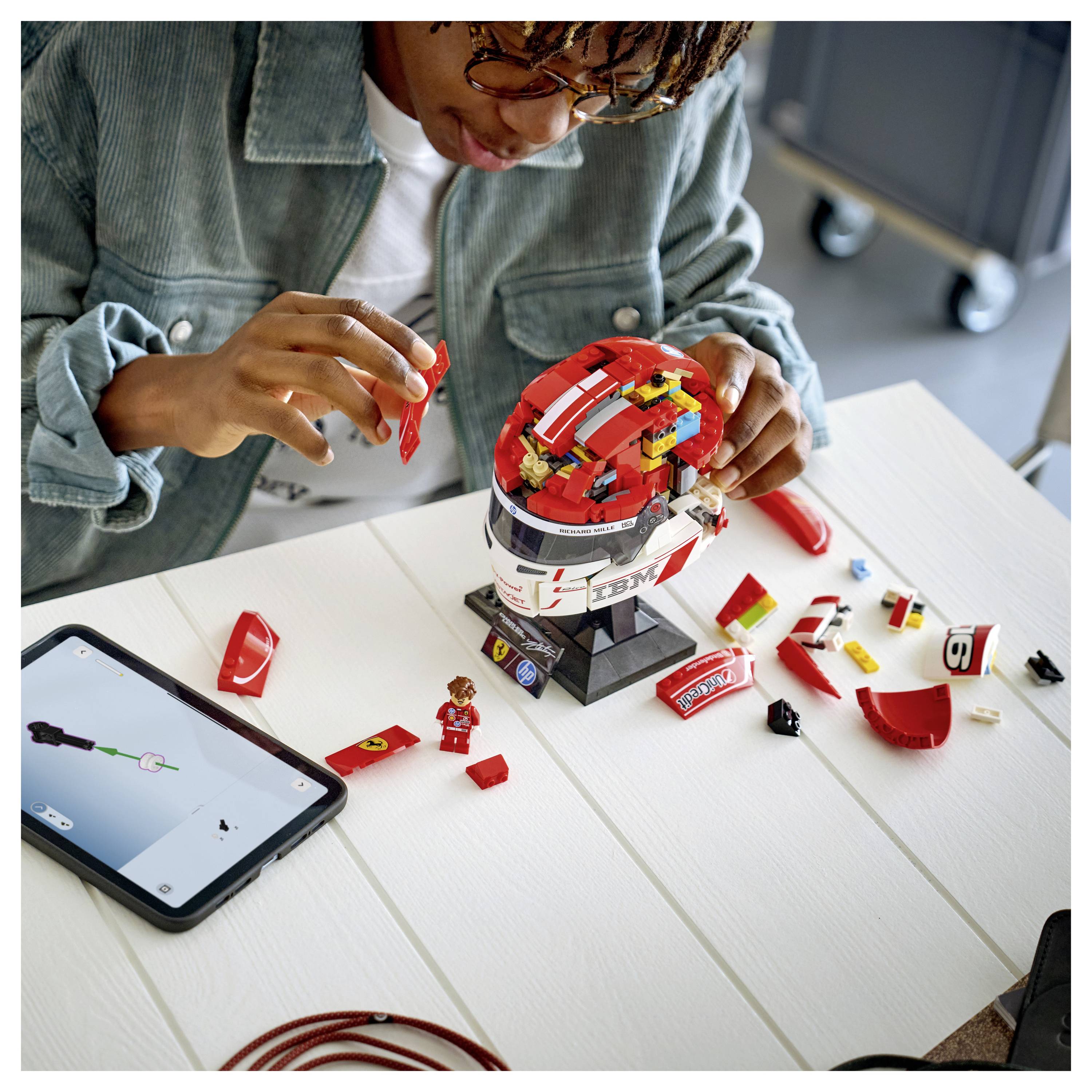 Eine Person setzt ein rotes LEGO-Helmmodell an einem Tisch zusammen, während sie eine digitale Anleitung auf einem in der Nähe positionierten Tablet verwendet.