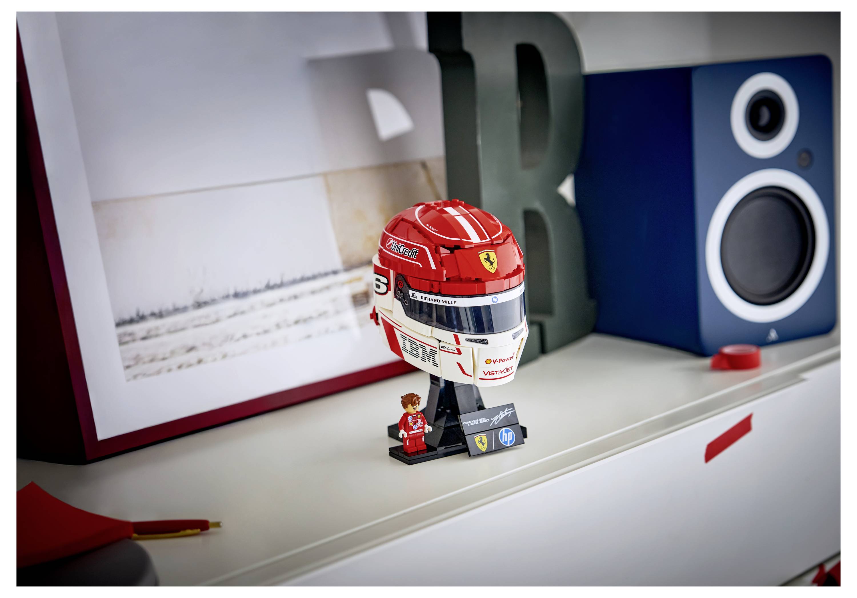 Ein detailliertes Spielzeughelm mit Ferrari-Logo und der Nummer 5 sitzt auf einem Regal neben einer Figur, umgeben von fotografischer Kunst und einem Lautsprecher.
