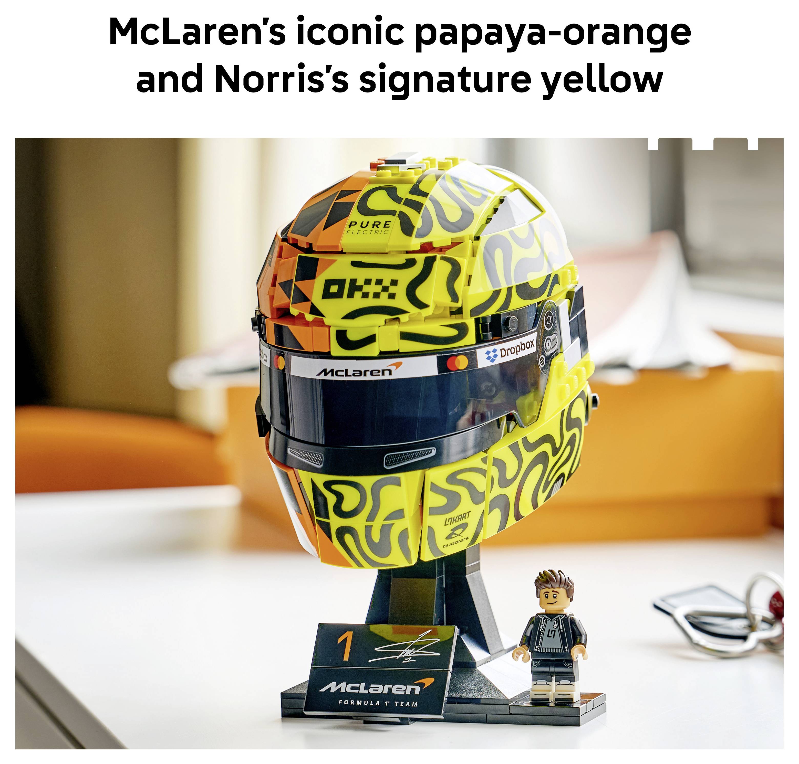 McLaren Formula-1-Helm in orange-gelber Lackierung auf Ausstellung, mit einer kleinen Minifigur und Teamplakette darunter.
