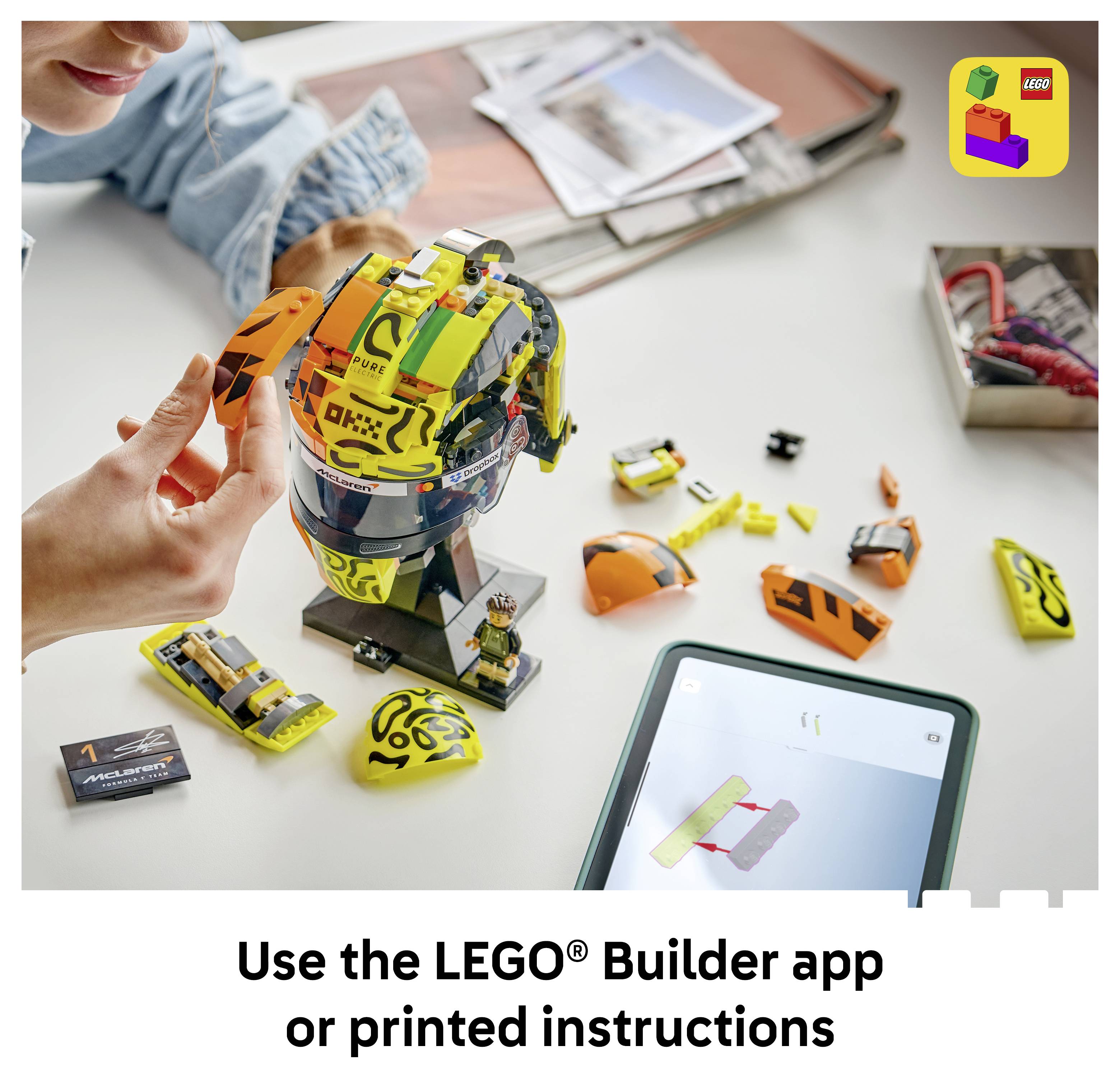Eine Person baut ein LEGO-Rennhelm-Modell auf einem Tisch zusammen, mit losen Teilen und einem Tablet, das Anweisungen anzeigt; der Text besagt „Verwenden Sie die LEGO® Builder-App oder gedruckte Anweisungen