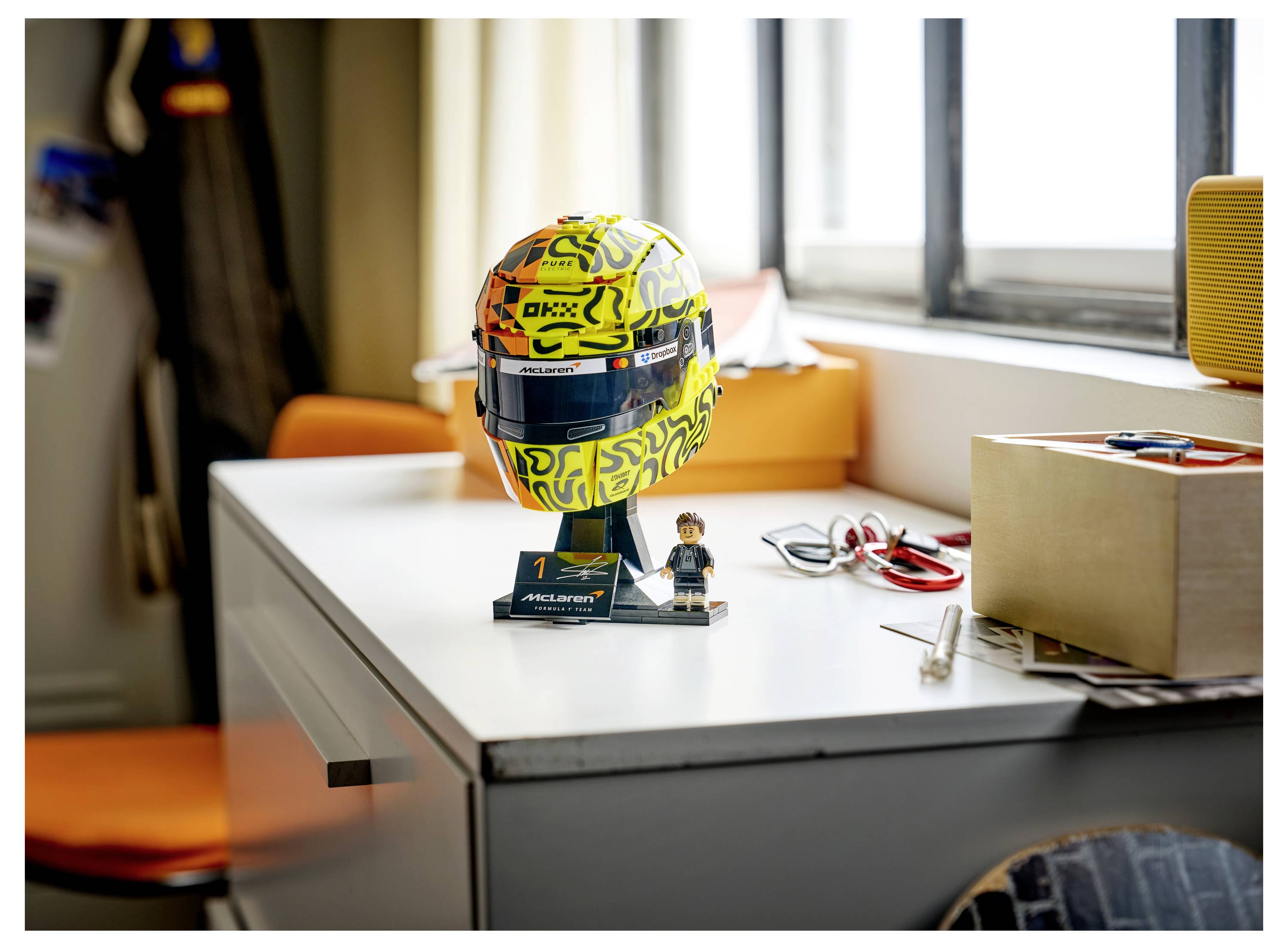Ein kleiner Modellrennhelm auf einem Schreibtisch mit einem McLaren-Namensschild und einer LEGO-Minifigur, neben Werkzeugen, Kabeln und einem Karton in der Nähe eines Fensters.