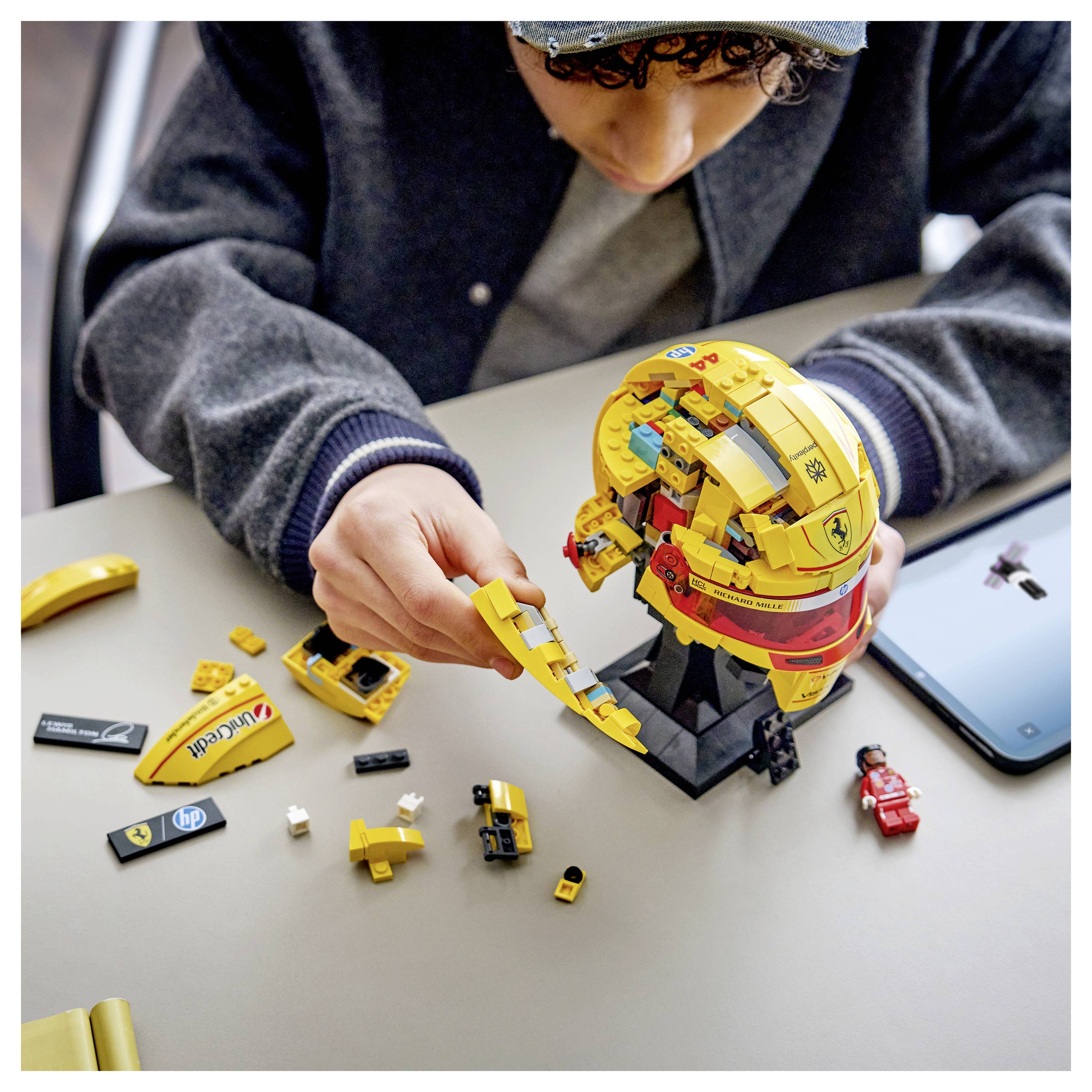 Eine Person setzt einen gelben LEGO-Globus auf einem Tisch zusammen, umgeben von LEGO-Steinen und einer roten Minifigur, was auf eine Bauaktivität hindeutet.