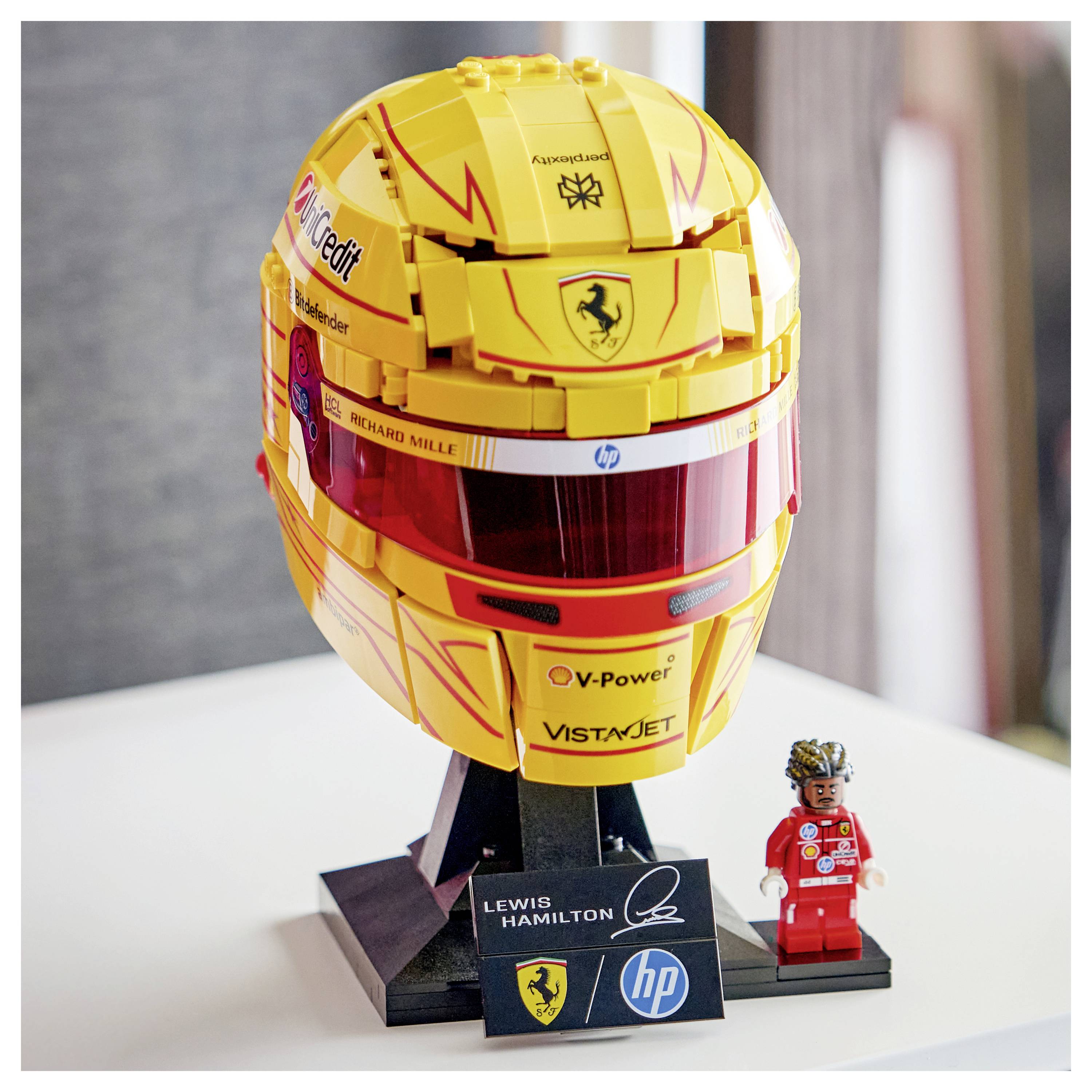 Ein gelber LEGO-Helm, der einem Rennhelm ähnelt, mit verschiedenen Sponsor-Logos. Daneben eine kleine LEGO-Figur in einem roten Rennanzug.