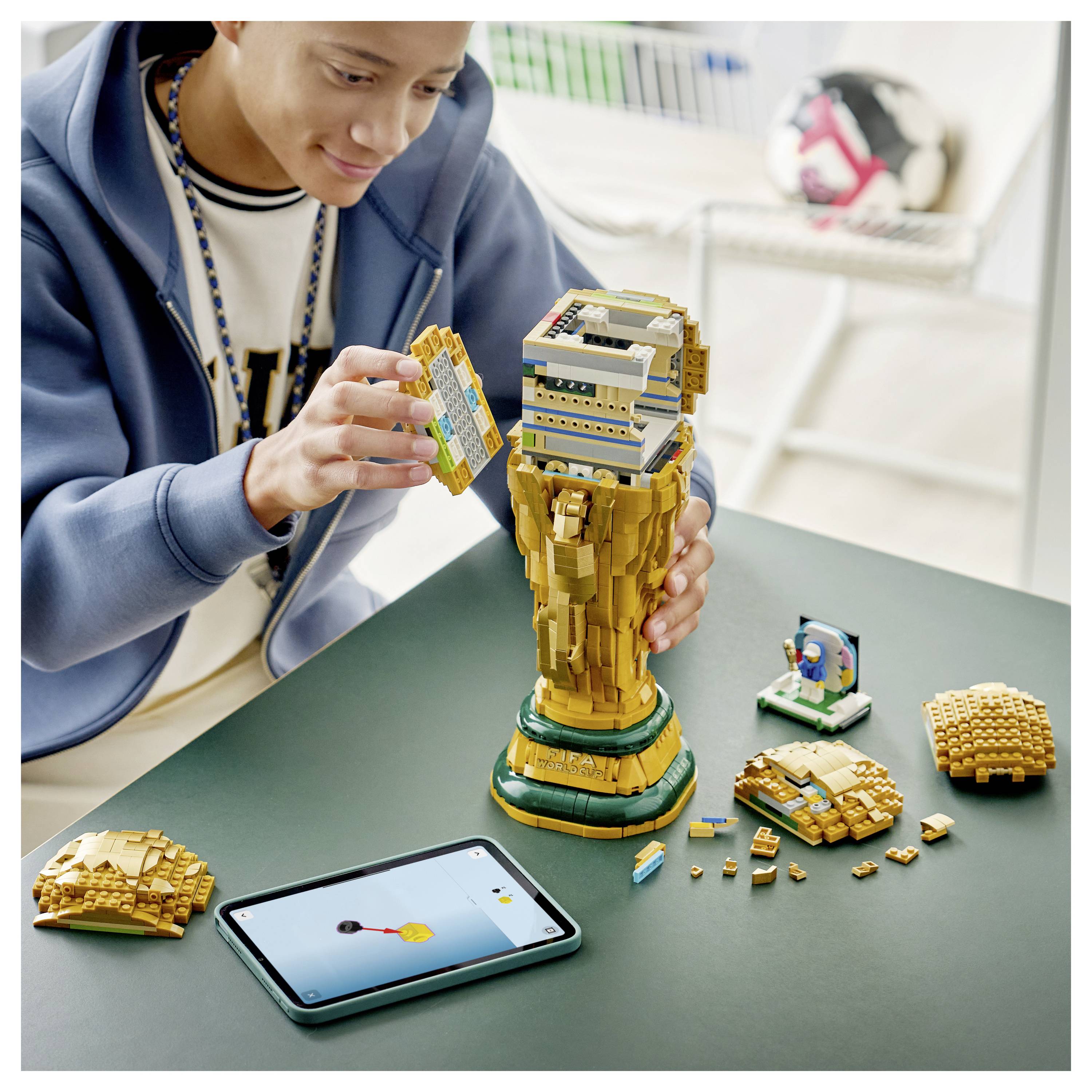 Eine Person baut ein hohes LEGO-Pokal-Modell auf einem Tisch zusammen, während auf einem Tablet eine Animation angezeigt wird. Verschiedene LEGO-Steine liegen verstreut in der Nähe.